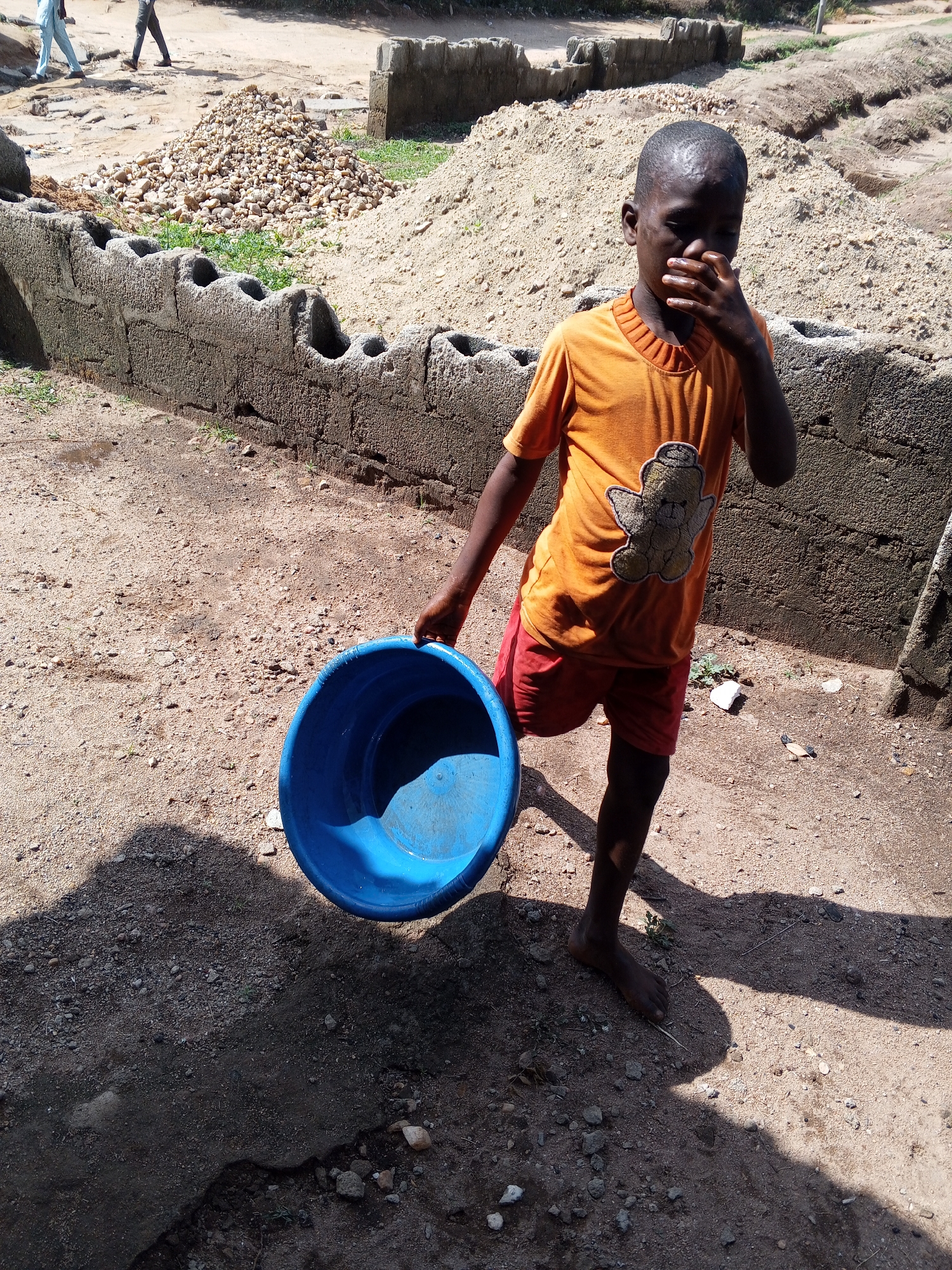 In Adonkolo, Nigeria, a young boy stands on bare ground in front of a low, uneven concrete block wall. He is wearing an orange t-shirt with a cartoon bear and red shorts. He holds a blue plastic bucket in his left hand and covers his mouth with his right. Behind him are piles of gravel and sand. In the background, two figures are walking away, and nearby, there are other low-lying concrete structures. The ground appears dry and dusty, with visible shadows.