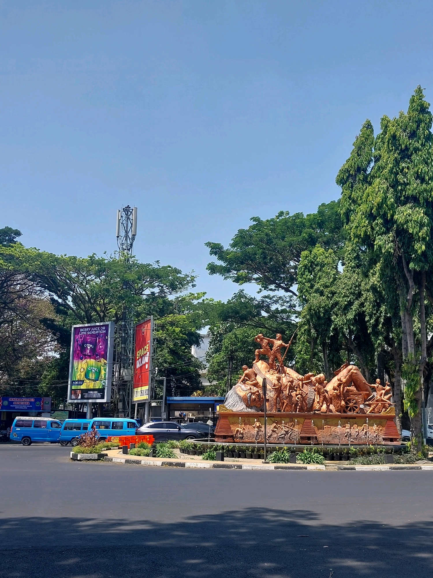 This is an outdoor, daytime, eye-level, vertical-format photograph taken on a clear day in the city of Malang, Indonesia. The main subject is a large, tiered bronze-colored monument situated in the center of a roundabout. The monument depicts numerous figures in various poses, some appearing to be engaged in labor or conflict, with detailed bas-relief sculptures around its base.
Surrounding the roundabout are several elements of an urban environment. To the left, there are three blue vans and a dark grey SUV. Behind the vehicles, a tall cell tower with multiple antennas is visible, with large billboards next to it. One billboard features an advertisement for Camel cigarettes and berry juice, while another displays the word "SPACE". A sign with the words "SENTRA KULINER SHIJDAYA" is also visible.
Lush green trees, some tall and slender and others with wide canopies, form a dense backdrop behind the monument and surrounding buildings. The sky is a clear, light blue. The road in the foreground is asphalt, with painted lane markers and shadows cast from trees.