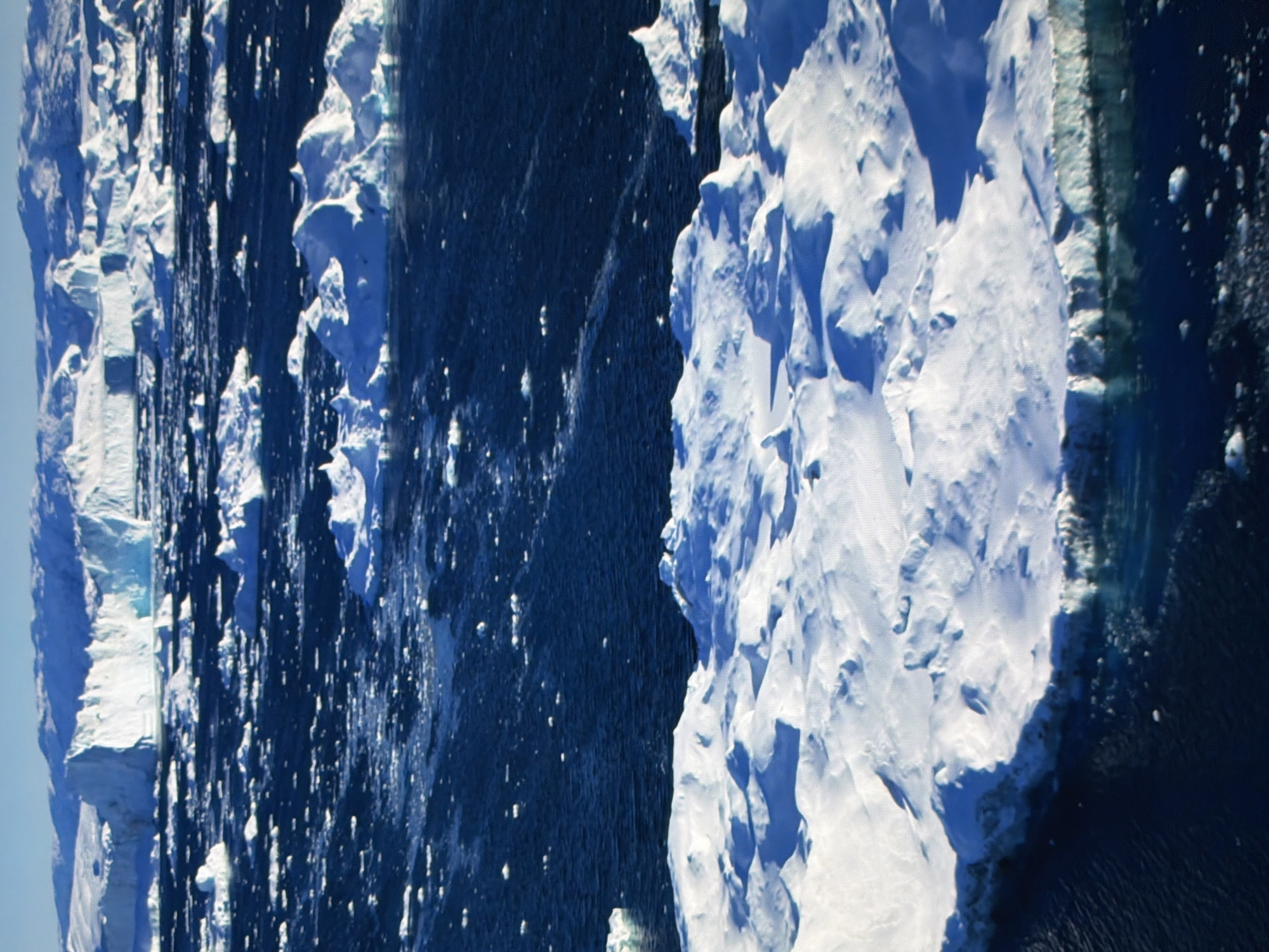 This aerial image captures a dramatic scene of icebergs and frigid ocean water. The dominant subjects are the large, jagged ice formations, characterized by their stark white surfaces and rugged textures, contrasting sharply with the deep, dark blue of the surrounding sea. The water is dotted with smaller pieces of ice, creating a textured surface.
The image appears to depict an environment in a polar region, likely Antarctica or the Arctic, given the presence of massive icebergs and the cold, rough ocean. The time of day seems to be during daylight hours, as indicated by the shadows and the overall illumination of the ice. The weather appears to be clear but cold, typical of such an environment.
There are no people or man-made objects visible in the frame. The scale of the icebergs suggests a vast, natural landscape. The mood evoked is one of raw, untamed nature and the power of the elements. The sharp, angular forms of the ice convey a sense of a formidable and potentially hazardous environment.
The image is a still photograph and does not show any activity or event taking place beyond the natural processes of ice formation and movement in the ocean. There is no visible text in the image. The provided location context of Kyiv, Ukraine, is inconsistent with the visual evidence in the image, which strongly suggests a polar environment.
