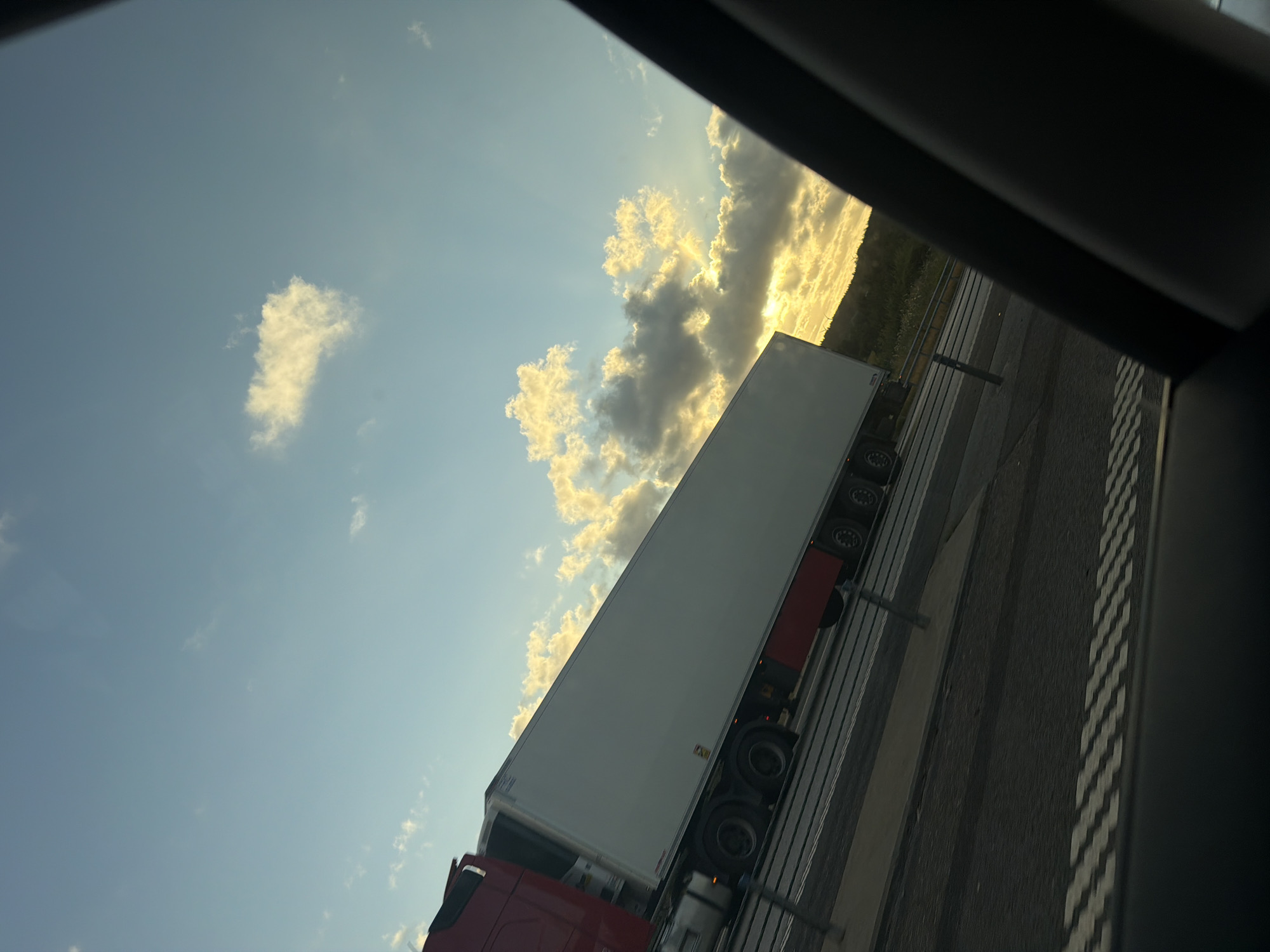 This is an image of a semi-trailer truck on a highway. The truck is red and white and is carrying a white trailer. The truck is moving from left to right. The sky is blue with some clouds. The sun is setting, and the light is coming from the right. The image is taken from inside a car.
The specific location in the city of Uddevalla, Sweden is not identifiable from the image.