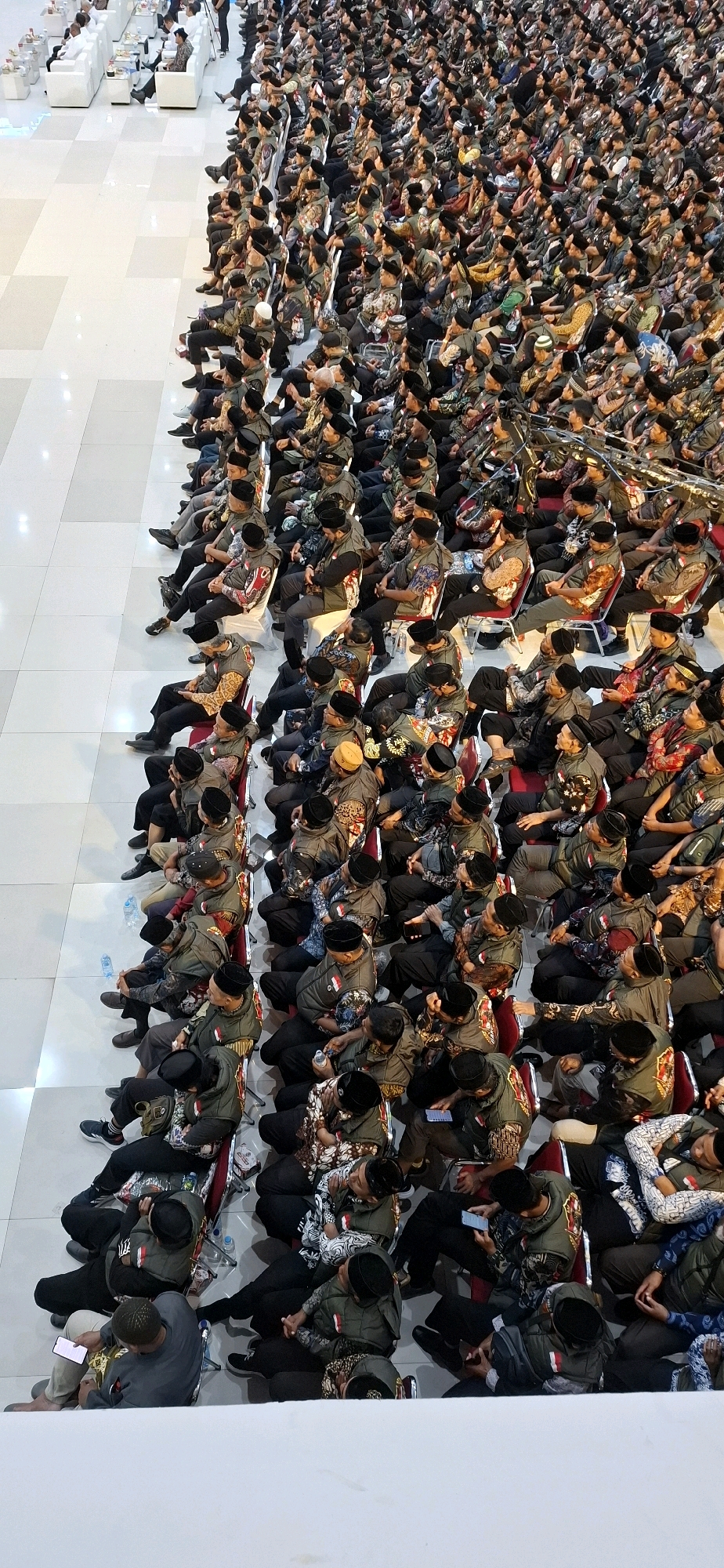 An elevated, high-angle perspective captures a large assembly of people seated in rows within what appears to be an indoor venue. The individuals are predominantly male, many wearing dark-colored caps, and some are adorned in matching olive-green vests over their clothing, suggesting a uniformed group. The seating arrangement consists of chairs, some red and some white, arranged in neat rows. The floor is a light-colored tiled surface, with a wider, lighter expanse to the left of the seated crowd, potentially a walkway or open area. In the background, to the far left, is a section with white furniture, including couches and chairs, occupied by a smaller group of individuals. The overall impression is one of an organized gathering or event taking place in a spacious hall, possibly a conference center or similar venue. The presence of bottled water on the floor indicates the duration of the event. The scene is static, with most individuals seated and some looking down or forward, suggesting they are listening to a speaker or observing something in front of them. There is no indication of the specific location within Blulukan, Indonesia, based solely on the visual cues in the image.