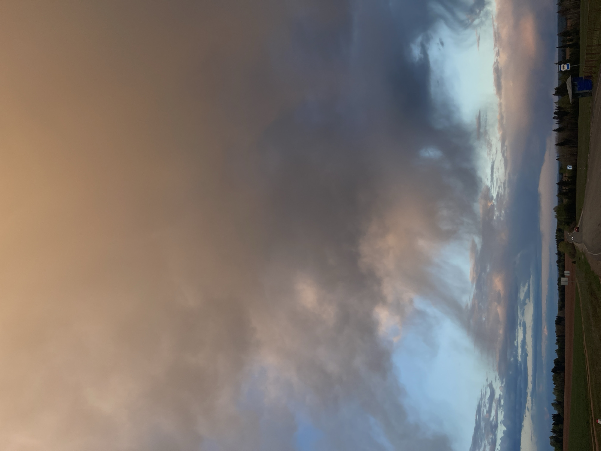 The image shows a cloudy sky over the location in the city of Перевозное, Russia. The sky transitions from an orange hue on the left to shades of blue and gray. The clouds appear thick and varied in density. A strip of land with trees and a road runs along the right side of the image. There is what looks to be a parking area. There are no visible people.