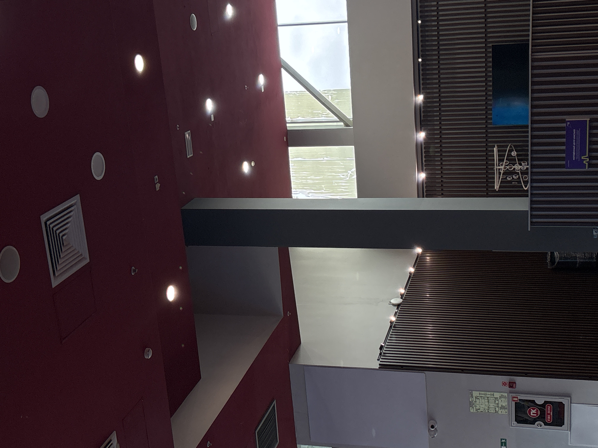 The image captures an interior space in Perm, Russia, likely a shopping or commercial center, characterized by a multi-story architectural design. The color palette is dominated by a deep red-burgundy hue on one wall, contrasting with neutral shades of white and gray used in other sections. Artificial lighting fixtures, including ceiling-mounted circular lights and linear lights along the ceiling's edge, illuminate the area. Architectural elements include a large pillar bisecting the view, skylights allowing natural light to filter in, ventilation grills, and window fixtures. Signage with emergency evacuation plans and fire hose details are visible on one wall. The ceiling features a wood-slat design with decorative elements. The space seems devoid of people and the general ambiance is modern.