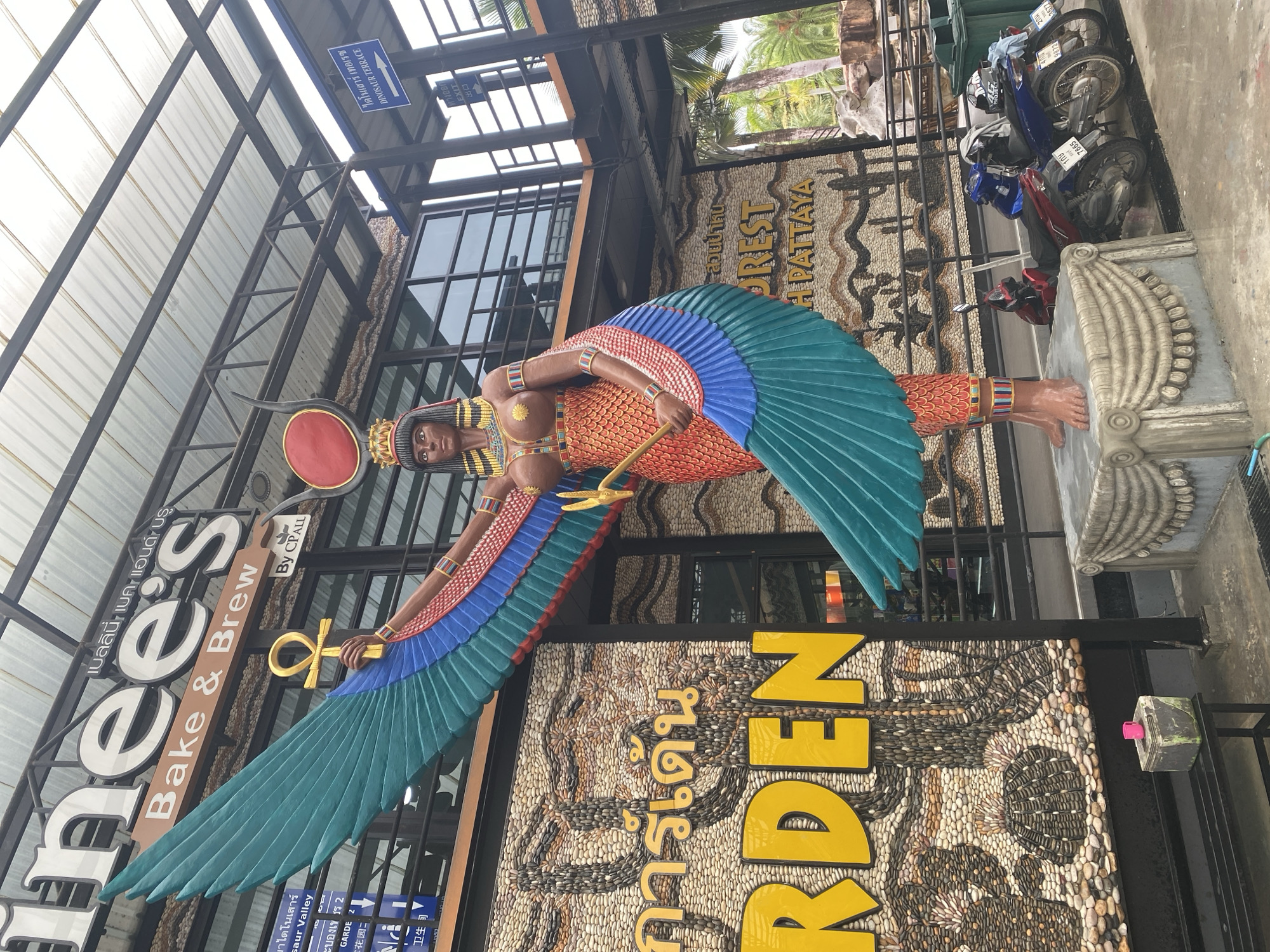 In Tambon Na Chom Thian, Thailand, a large, colorful statue of a winged Egyptian deity stands prominently in front of a building. The statue, adorned with a red headdress and golden jewelry, holds a golden ankh. Its wings are a vibrant mix of teal, blue, and red. Behind the statue, the building features signage in English and Thai, with "Bake & Brew" clearly visible. The wall behind the statue is a mosaic of pebbles, depicting a face and tropical plants. To the right of the statue, several motorcycles are parked, and a green trash bin is visible. Palm trees and other vegetation suggest a tropical environment. The ground is paved, and there is a black bench in the foreground.