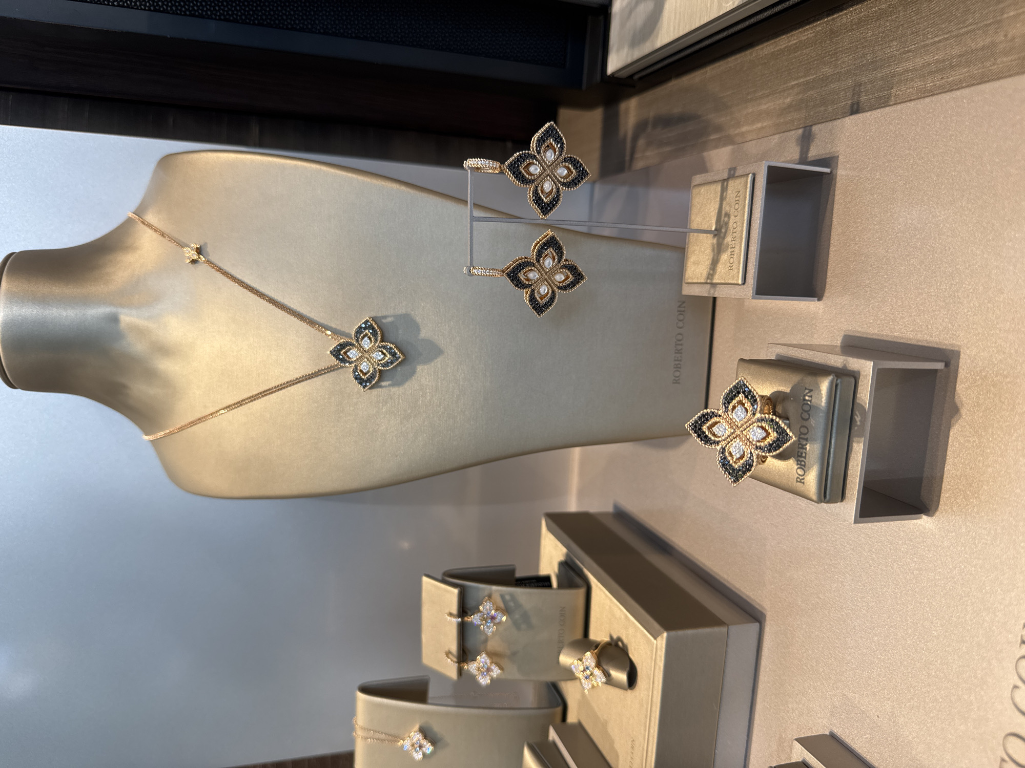 An image displays a collection of Roberto Coin jewelry arranged in a store display. The jewelry, primarily necklaces, earrings, and rings, is presented on a mannequin bust, stands, and display boxes. The pieces feature intricate designs with diamonds and dark stones, likely sapphires. The overall aesthetic is luxurious and elegant, with soft lighting highlighting the precious materials. The store's branding, "Roberto Coin," is visible on several display stands. The image is shot from a slightly elevated angle, capturing the arrangement from left to right. The background is a neutral, light-colored wall, suggesting an indoor retail environment.