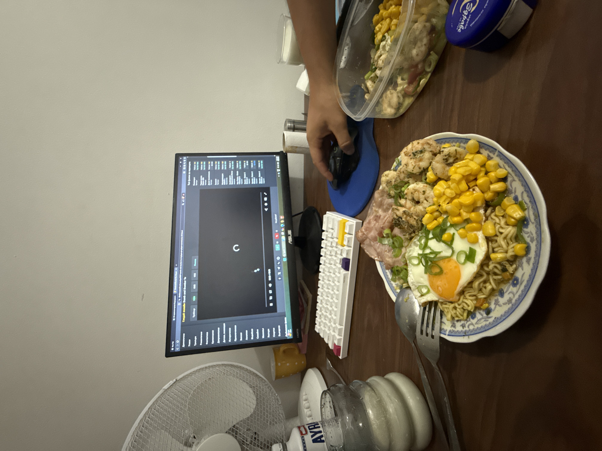 The image depicts a desk scene with a computer monitor displaying a video streaming service. A person's hand is visible on a computer mouse, interacting with the screen. On the desk, there is a plate of food containing noodles, a fried egg, corn, and meat. A container with a salad is also present, along with a glass of milk and a small blue tin. A white keyboard and a fan are also part of the setup. The background shows a plain wall. There is no information in the image to suggest the location is in Rückingen, Germany.