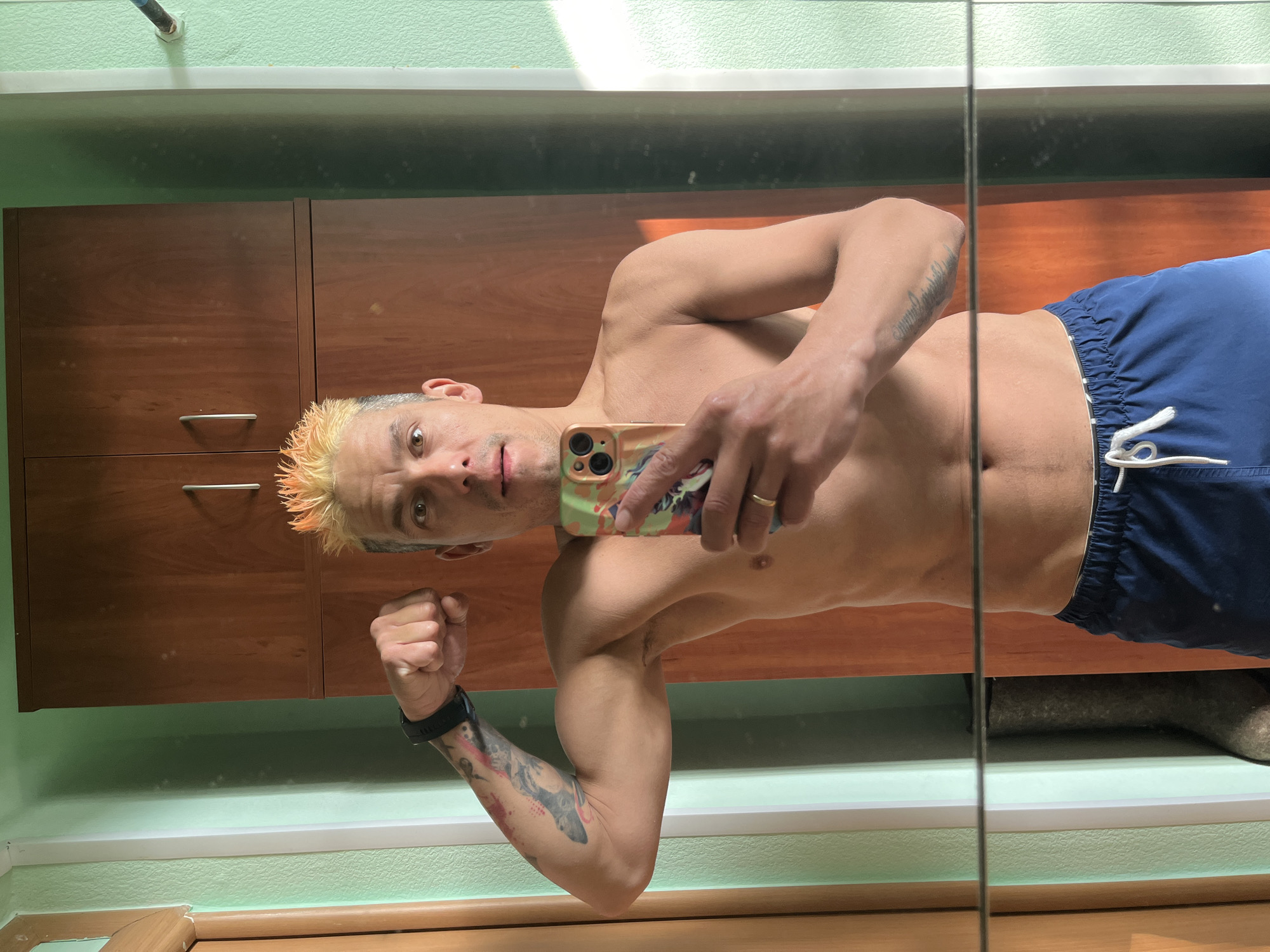A shirtless man with dyed orange hair is taking a selfie in a mirror. He is wearing blue swim trunks and a black smartwatch. He is flexing his left bicep. The background appears to be a bathroom, with wooden cabinets and light green walls. The image might be located in the city of Chaikovsky, Russia, due to the user's request, but that is not discernable from the image itself.