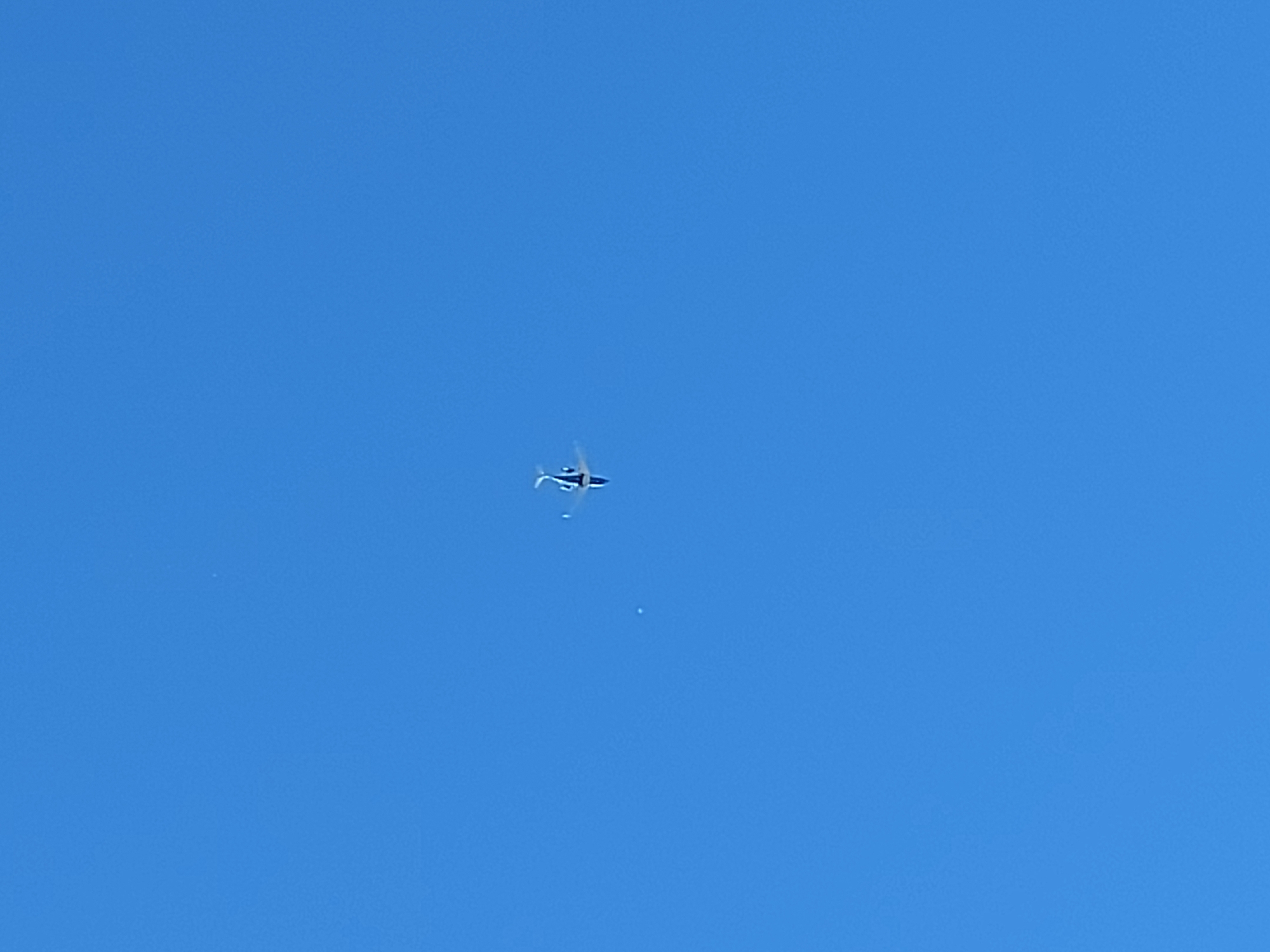 A single white helicopter with black accents is visible in the upper-middle section of the image. The helicopter appears to be in flight against a clear blue sky. There are two small, faint, white circular shapes or smudges in the sky to the left of the helicopter. The environment is a bright, cloudless sky, suggesting an outdoor daytime scene. There are no visible landmarks or indications of the city of Calgary, Canada.