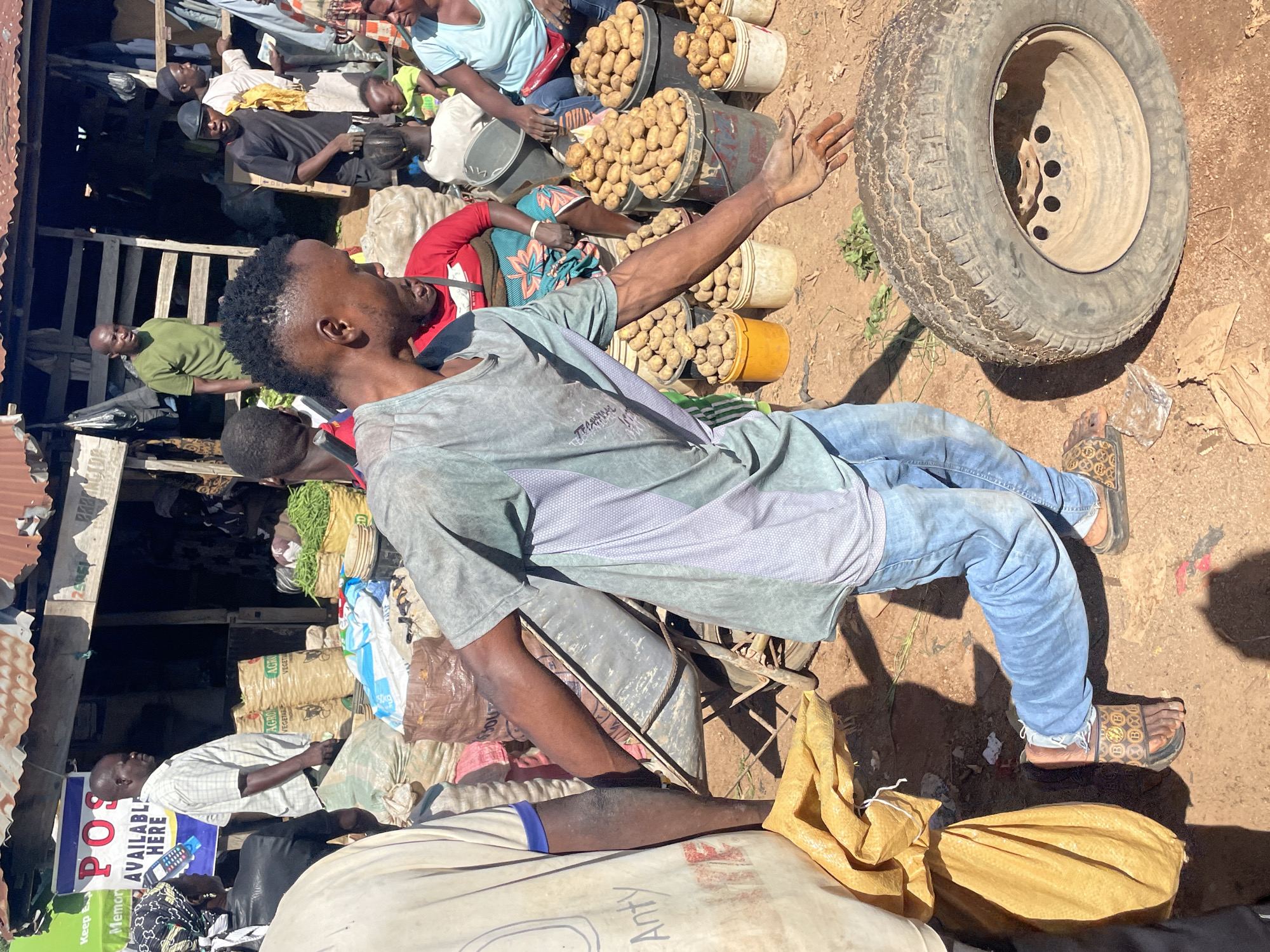 In Gura Top, Nigeria, a bustling marketplace scene unfolds under a bright sun. A man, his face turned away from the camera, is the central figure, dressed in a grey t-shirt and blue jeans, with his left arm extended towards a large tire lying on the dusty ground. He is in motion, his body leaning forward as if about to interact with the tire. To his left, another man, wearing a plaid shirt, stands with his hands clasped, looking to the side. Behind him, a stall displays various sacks of produce, including what appear to be potatoes and green beans, suggesting a vibrant agricultural trade.
Several other individuals are present in the background. A woman in a bright red top and patterned skirt is seated, her attention seemingly directed elsewhere. Further back, a woman in a light blue top is visible, and a child in a green garment can be seen. The environment is characterized by a makeshift structure with a corrugated metal roof, common in local markets. The ground is dirt, and the overall impression is one of lively commerce and daily life in the community.