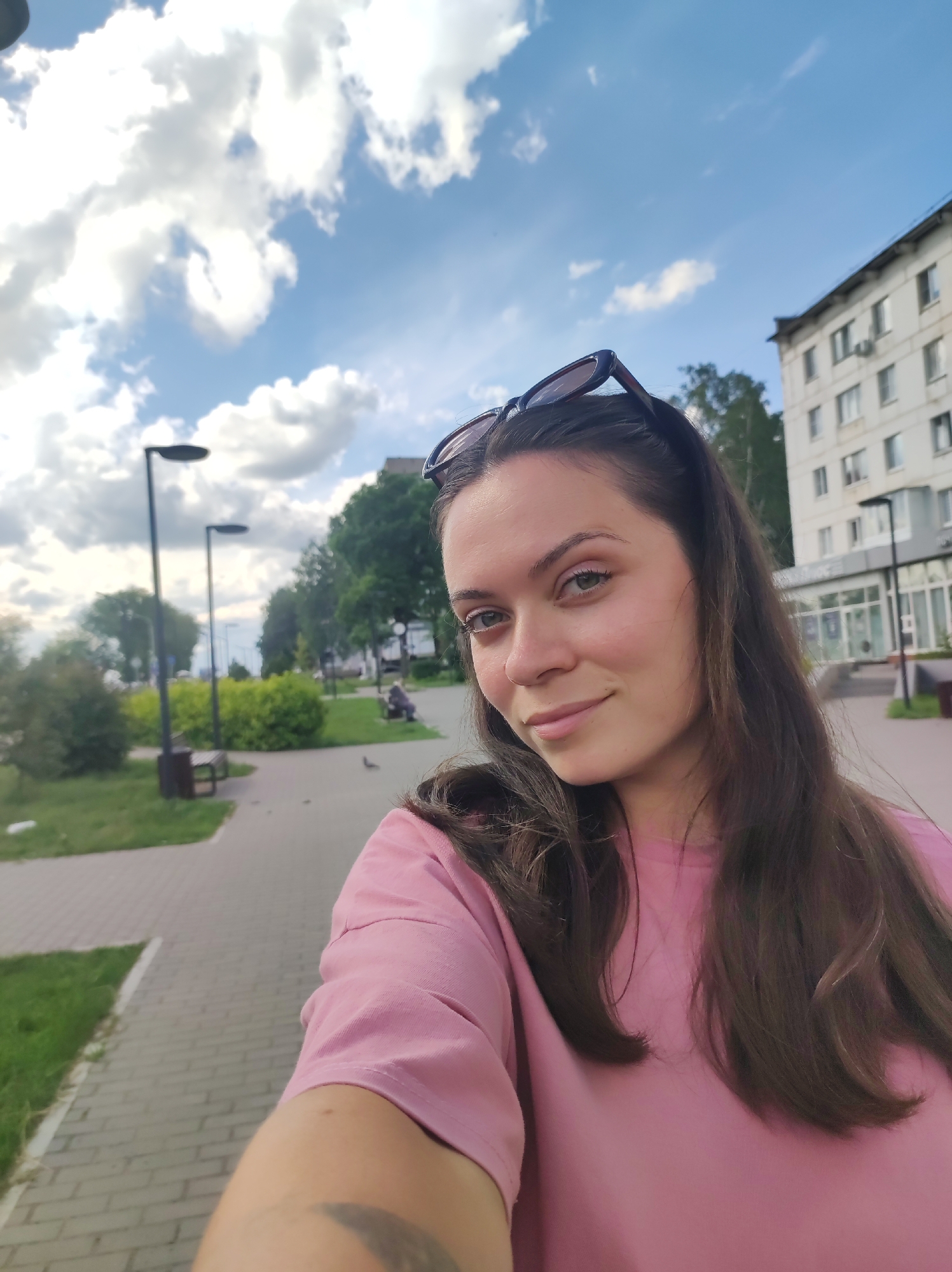 A young woman with dark brown hair and light green eyes is taking a selfie outdoors in Chaikovsky, Russia. She is wearing a pink t-shirt and has sunglasses on top of her head. She is smiling and looking at the camera. The background shows a paved walkway, a grassy area with benches and lampposts, trees, and a multi-story building. The sky is blue with white clouds.