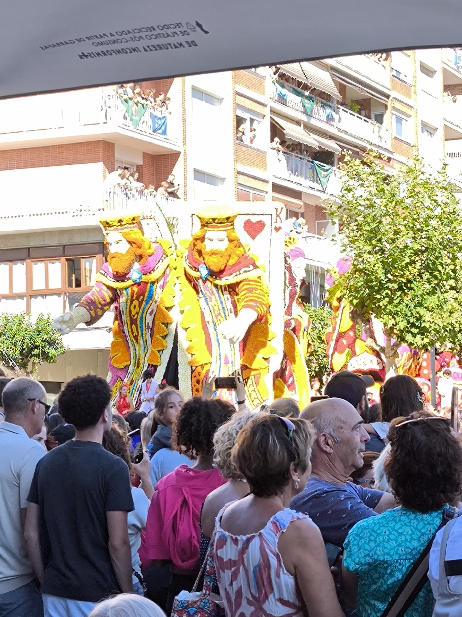 This photograph captures a daytime scene in the city of Laredo, Spain, likely during a festival or parade. In the foreground, a diverse crowd of people is visible, with individuals looking in various directions, some appearing to be observing an event. Their clothing is casual and varied, suggesting a public gathering.
Dominating the mid-ground are two large, elaborate floral sculptures depicting figures dressed as kings or royalty. These figures are intricately decorated with vibrant flowers, showcasing detailed craftsmanship. One sculpture prominently features a heart symbol and the letter "K," hinting at a possible playing card theme. Behind the floral sculptures, a multi-story building with balconies is visible, with people observing from the balconies. The architecture of the building suggests a European urban setting.
To the right of the floral sculptures, a leafy green tree adds a natural element to the scene. The overall atmosphere appears festive and lively, with a sense of community engagement. The image's perspective suggests it was taken from within the crowd, looking towards the main spectacle.