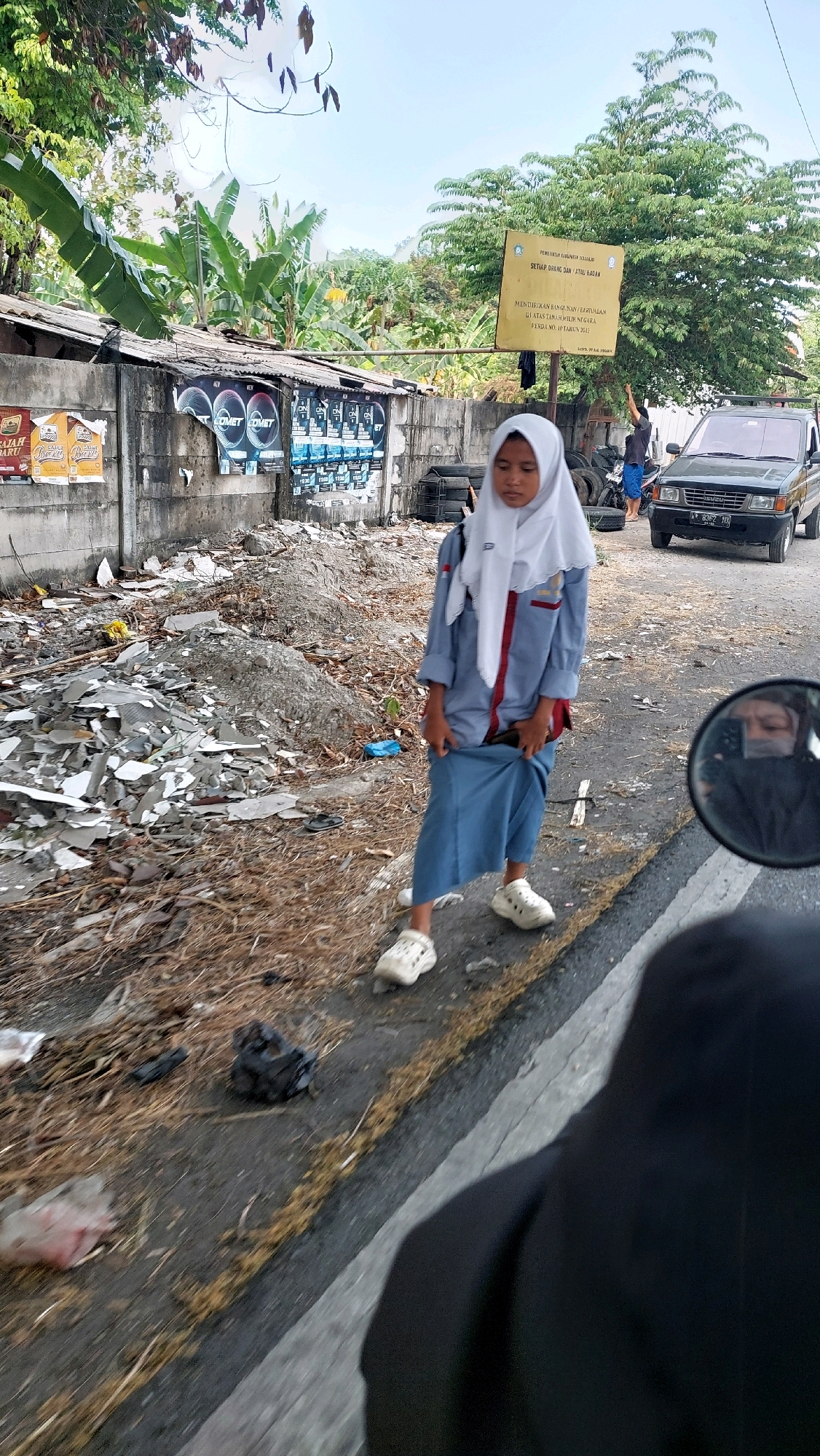 This media file captures a young person in a school uniform standing on a roadside in what appears to be Sidoarjo, Indonesia. The individual, wearing a white hijab and a light blue and grey uniform with red accents, holds the hem of their blue skirt. They are dressed in white clogs.
The scene is set on a sunlit day, suggested by the bright lighting and clear sky visible in the background. The setting is somewhat dilapidated, with a concrete wall covered in posters and debris piled up along the roadside. A signpost with Indonesian text is visible behind the person, indicating a public notice. Tires are stacked near the wall.
Further down the road, a dark-colored SUV is parked, and another person can be seen in the distance. The foreground shows the edge of a motorcycle's rearview mirror, reflecting the face of the person taking the photo. The overall impression is a moment captured in a daily urban or semi-urban environment, possibly during a school day. The subject's expression is neutral or slightly downcast, and their stance suggests they might be waiting or have paused briefly. The presence of trash and the weathered appearance of the wall hint at a less-than-pristine environment.
The visible text on the sign reads: "SETIAP ORANG DAN ATAU BADAN MENDIRIKAN BANGUNAN/BARTUALAN DI ATAS TANAH MILIK NEGARA PERDA NO. 10 TAHUN 2013". This translates to "Every person and/or body establishes buildings/structures on state-owned land, Regional Regulation No. 10 of 2013."