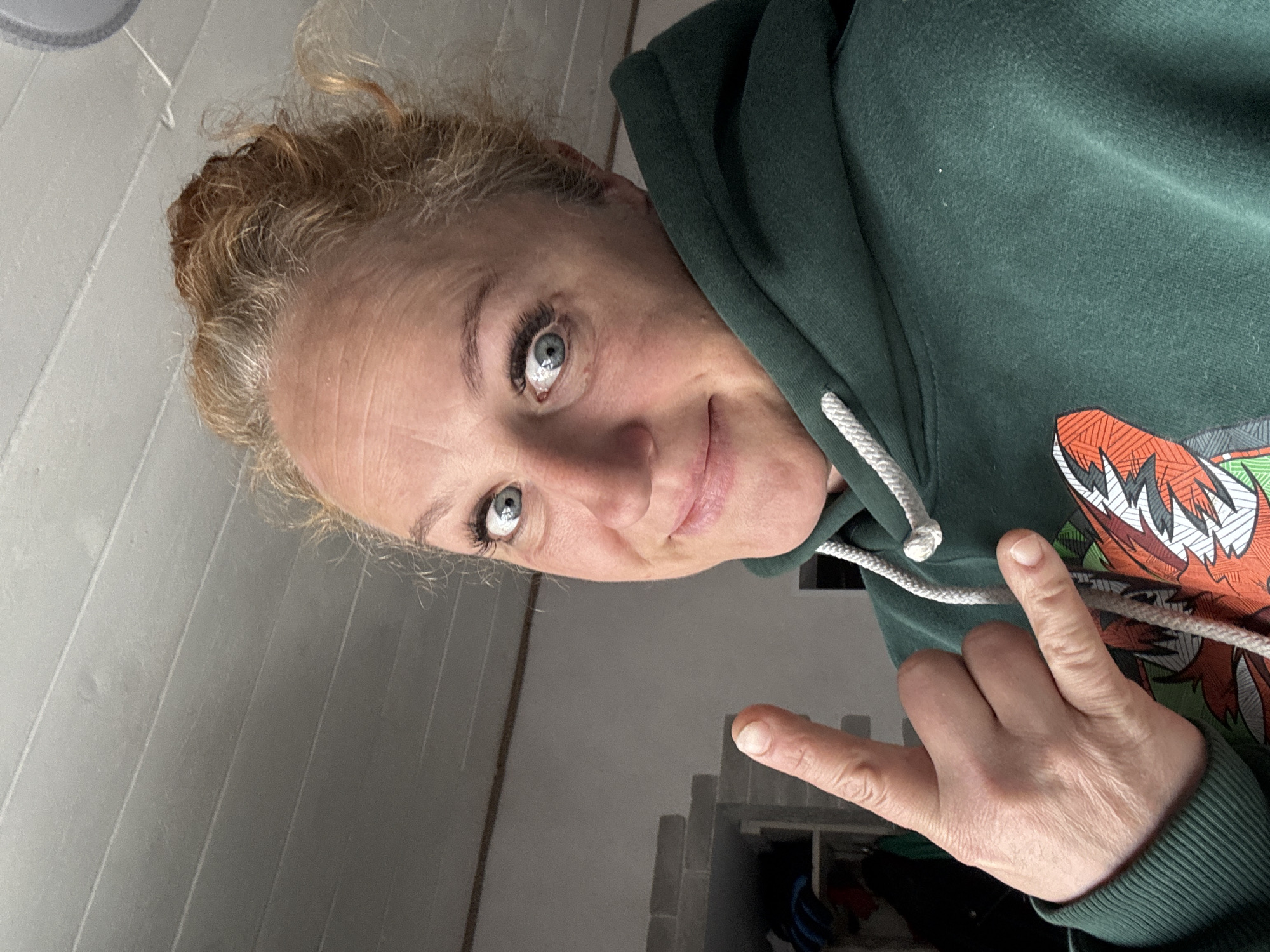 A selfie of a middle-aged white woman with blonde hair pulled back in a messy bun and blue eyes. She is wearing a dark green hooded sweatshirt with a graphic design on the chest. She is giving the "rock on" hand gesture. The background includes a gray paneled wall and what appears to be a storage area with stacked concrete blocks and possibly some items inside. The location is likely indoors, and no identifying information explicitly links the image to the city of Chaikovsky, Russia.