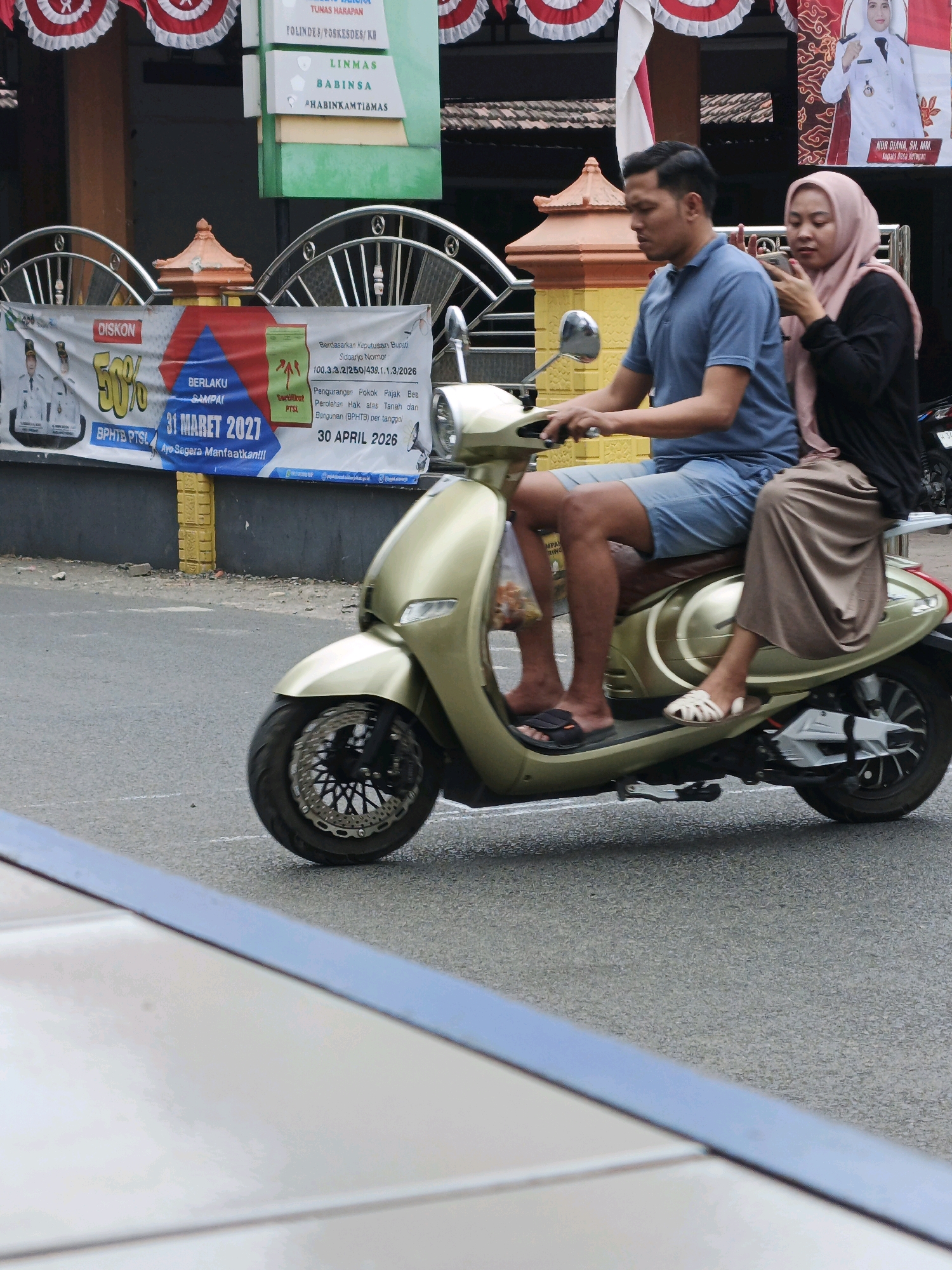 An Indonesian couple rides a gold scooter on a paved road in Ketegan, Indonesia. The man, driving, wears a blue polo shirt and denim shorts. The woman, riding on the back, wears a pink hijab and a long brown skirt, and holds a smartphone to her ear as if talking or texting. Both are barefoot. The scooter has a silver trim and a large, distinctive front wheel with a patterned disk brake. In the background, a banner with promotional text and images of government officials is visible, along with some red and white decorative banners. The environment suggests a street scene in a built-up area.
