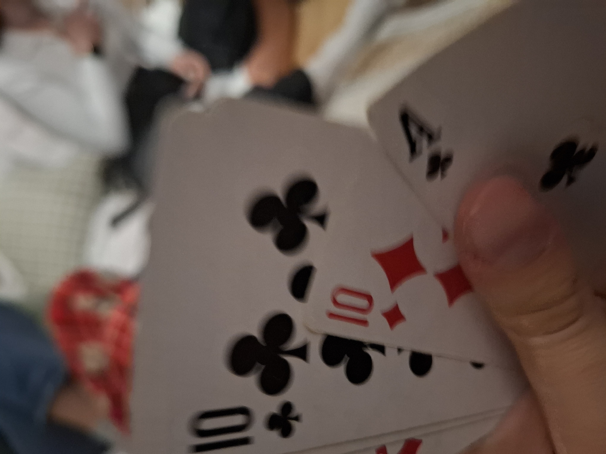 A hand holds a playing card, the 4 of Clubs, along with several other playing cards. One of the cards underneath is the 10 of Diamonds, and another is the 10 of Clubs. People are blurred in the background. The location, Panagyurishte, Bulgaria, cannot be determined from the visual information provided.