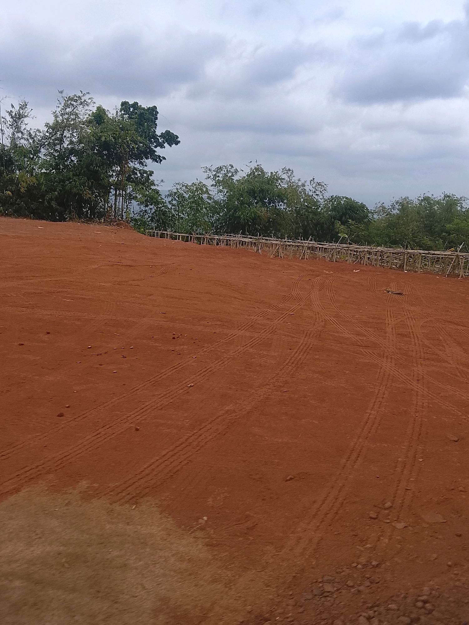 A wide shot of a dirt field with tire tracks on it, in Banjar, Indonesia. The field is red-brown and appears to be slightly sloped. In the background, there is a line of trees and a bamboo fence. The sky is cloudy.