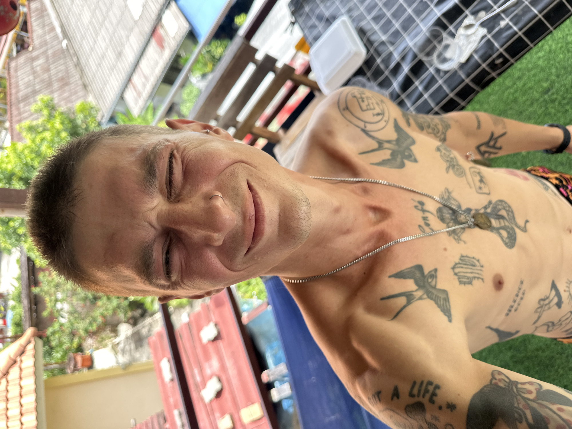 A topless man with short hair and tattoos on his chest and arms is taking a selfie outdoors in Karon, Thailand. He is winking and smiling. Behind him, a balcony overlooks rooftops and lush greenery. A table with a checkered cloth and a white container is also visible.