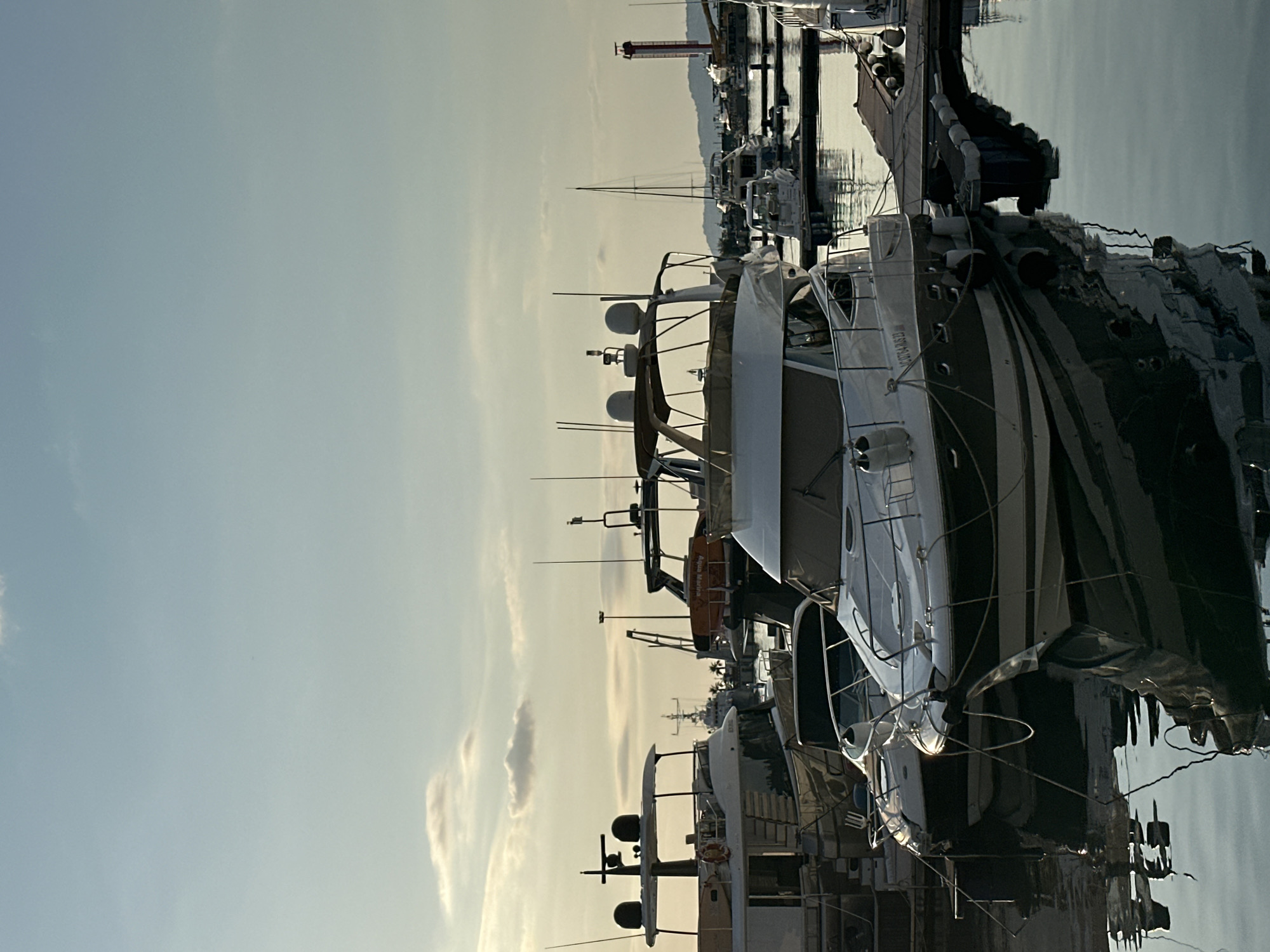 This is a photograph taken from a marina in Sochi, Russia. The image depicts a collection of yachts and boats moored in the water, with reflections visible on the calm surface. In the background, a red and white lighthouse stands tall on a hillside, with mountains and the sky visible beyond. The lighting suggests it may be late afternoon or early evening, with soft, warm light illuminating the scene. The sky is a pale blue with a few scattered clouds. The marina appears to be well-maintained, with wooden docks and mooring lines connecting the vessels. There are no people visible in the image.
