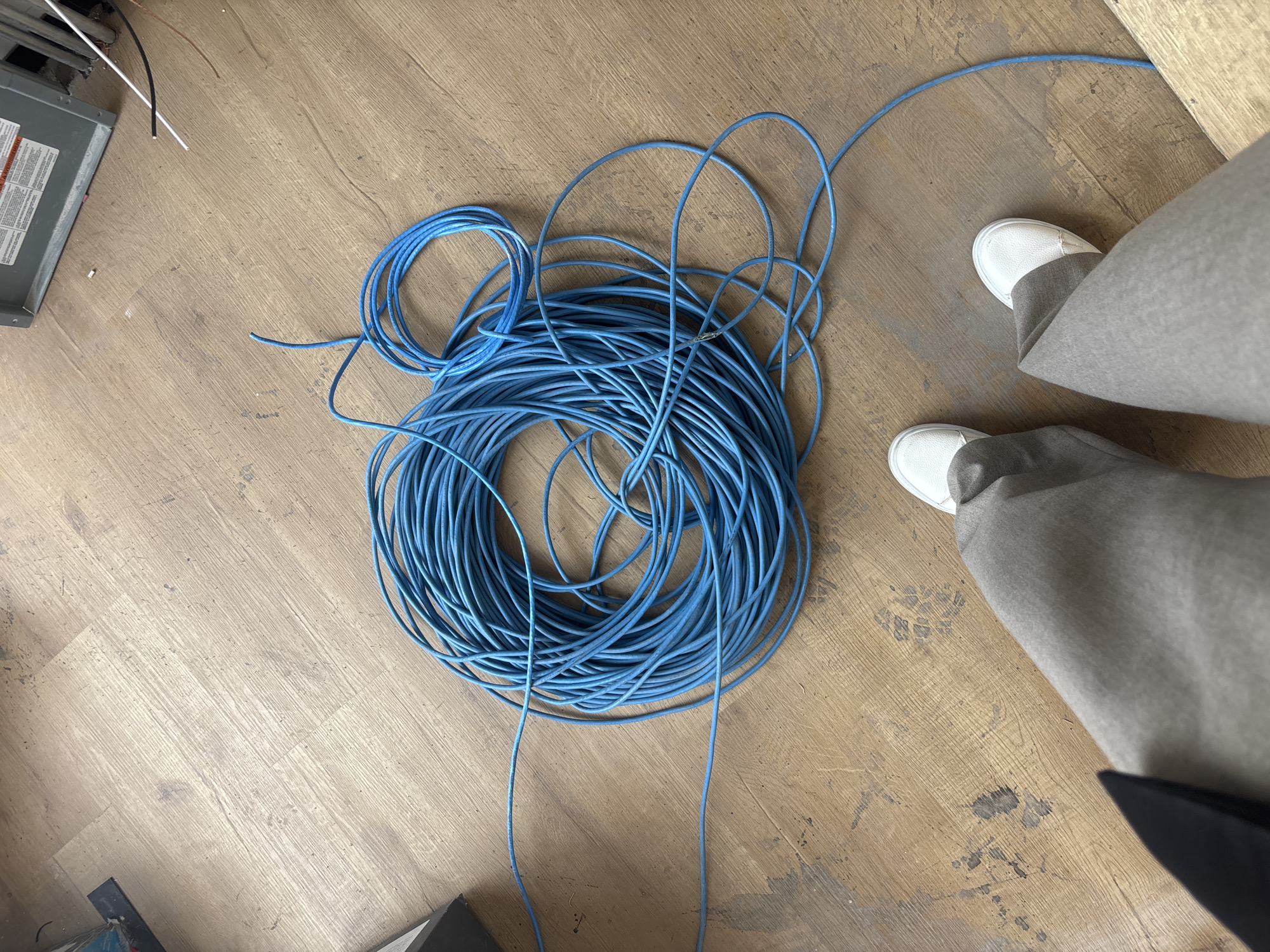 The image shows a coil of blue cable lying on a light brown wooden floor. The coil is somewhat disorganized, with some loops extending outwards. In the lower right corner, a person wearing gray pants and white shoes is standing. Parts of what appear to be electrical equipment are visible in the upper left and lower left corners of the image. The location in the city of Buenavista, Mexico is unknown.