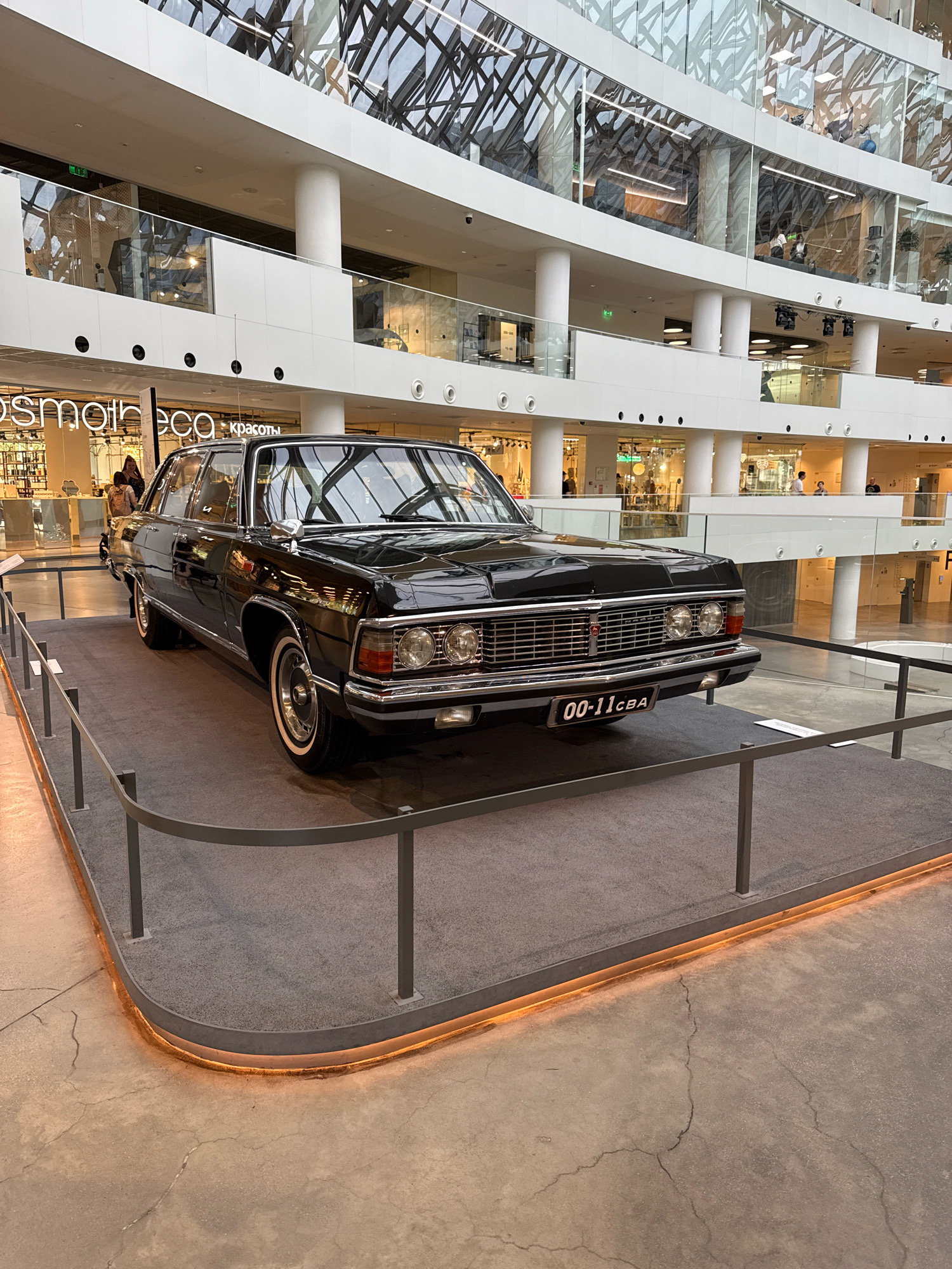 The image shows a black vintage GAZ-13 Chaika sedan displayed on a raised platform in a modern, multi-story shopping mall. The car is a centerpiece, positioned on a gray carpeted area surrounded by a metal railing. The mall's interior features glass railings, white columns, and a large, geometrically patterned glass ceiling that allows natural light to filter in. In the background, various shops are visible on different levels, with people browsing and walking. The license plate on the car reads "00-11 CBA." The scene appears to be a public space, likely a shopping center or exhibition hall, in Yekaterinburg, Russia, given the context provided.