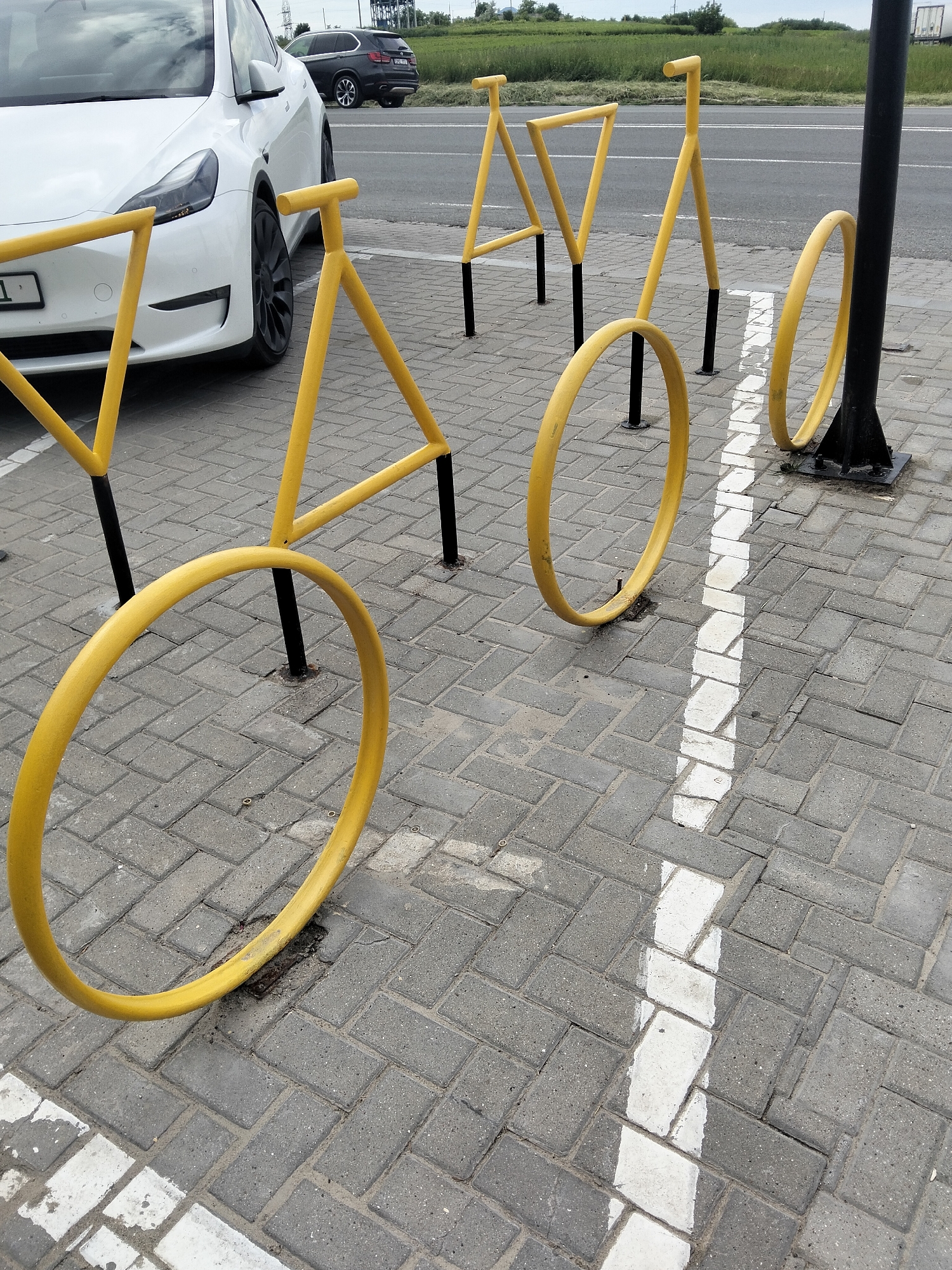 A white electric car is parked on a gray brick sidewalk. In front of the car are yellow bicycle racks shaped like bicycles. The racks are made of thick metal tubing and have black supports. There are white lines painted on the sidewalk, indicating parking spaces. In the background, a dark SUV is driving on a road, and beyond that, there are green fields and trees under a cloudy sky. The scene appears to be a parking lot or an area near a road. No people are visible. The overall impression is that of a functional outdoor space, likely a public area with facilities for both cars and bicycles. The time of day is daytime, and the weather is overcast. The location cues, such as the brick paving and the type of road visible, are consistent with Peresecina, Moldova. There is a license plate visible on the white car with the text "1".