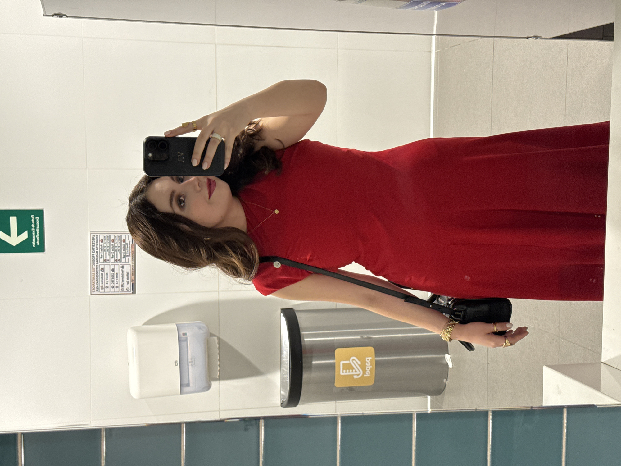 A woman stands in front of a bathroom mirror, taking a selfie. She is wearing a red dress and a black purse over her shoulder. Her dark hair is styled in waves. Rings adorn the fingers of both hands. She's holding a black smartphone. The bathroom is simple, with white tiles on the upper half of the walls and blue tiles on the lower half. There is a paper towel dispenser and a hand dryer mounted on the wall. A green exit sign is visible. Text is visible on the wall near the exit sign. The city of Mexico City, Mexico is not discernible.