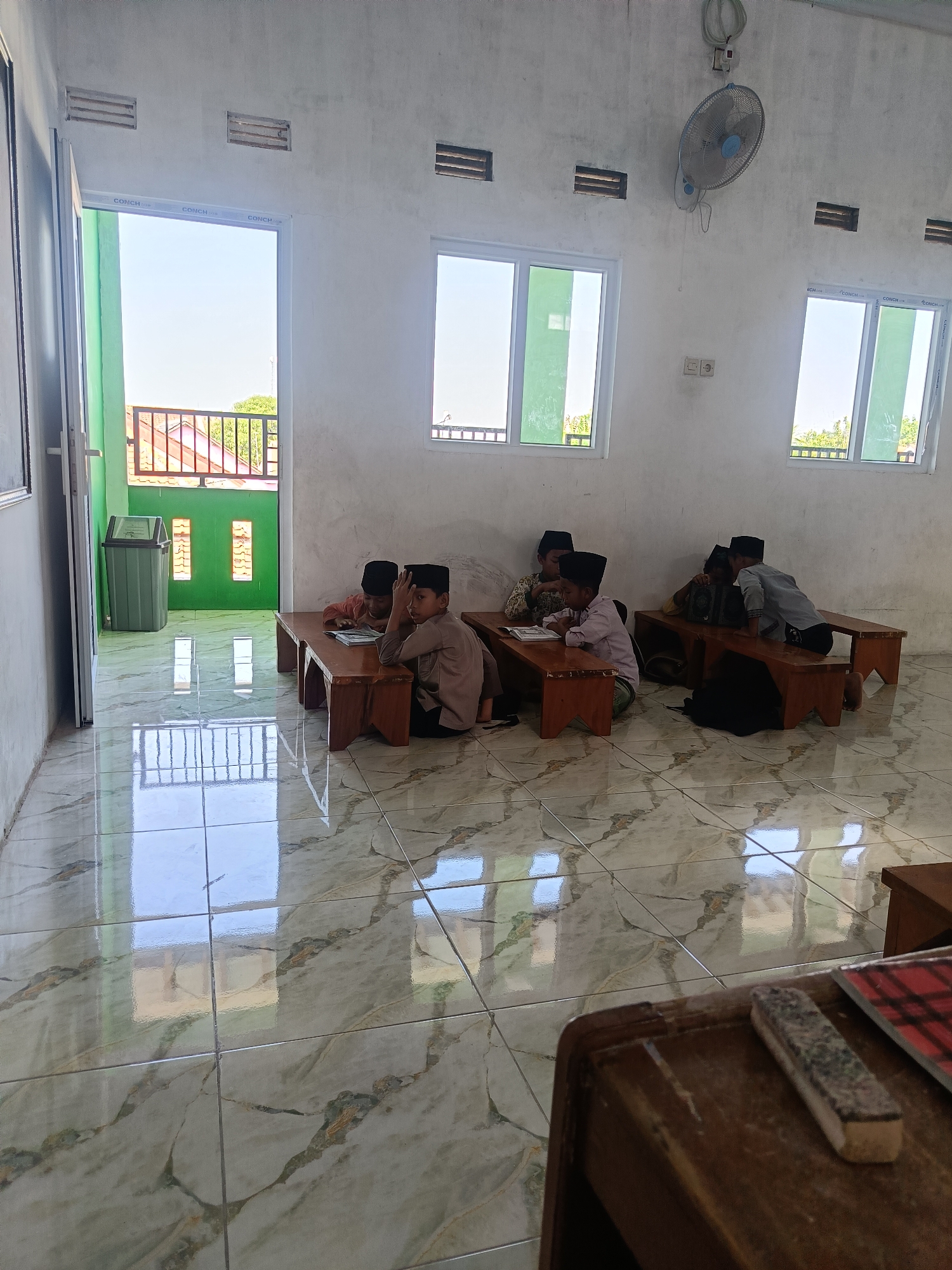 In a room in Pakijangan, Indonesia, five young boys, dressed in traditional Islamic attire, are seated at wooden benches, engaged in learning. The room has white walls and large windows that offer a view of the outdoors. A ceiling fan is mounted on the wall, and the floor is tiled with a marble-like pattern. In the foreground, a wooden desk is visible with a whiteboard eraser and a book. A doorway on the left leads to an outdoor area with a trash can. The overall atmosphere is one of quiet study and education.
