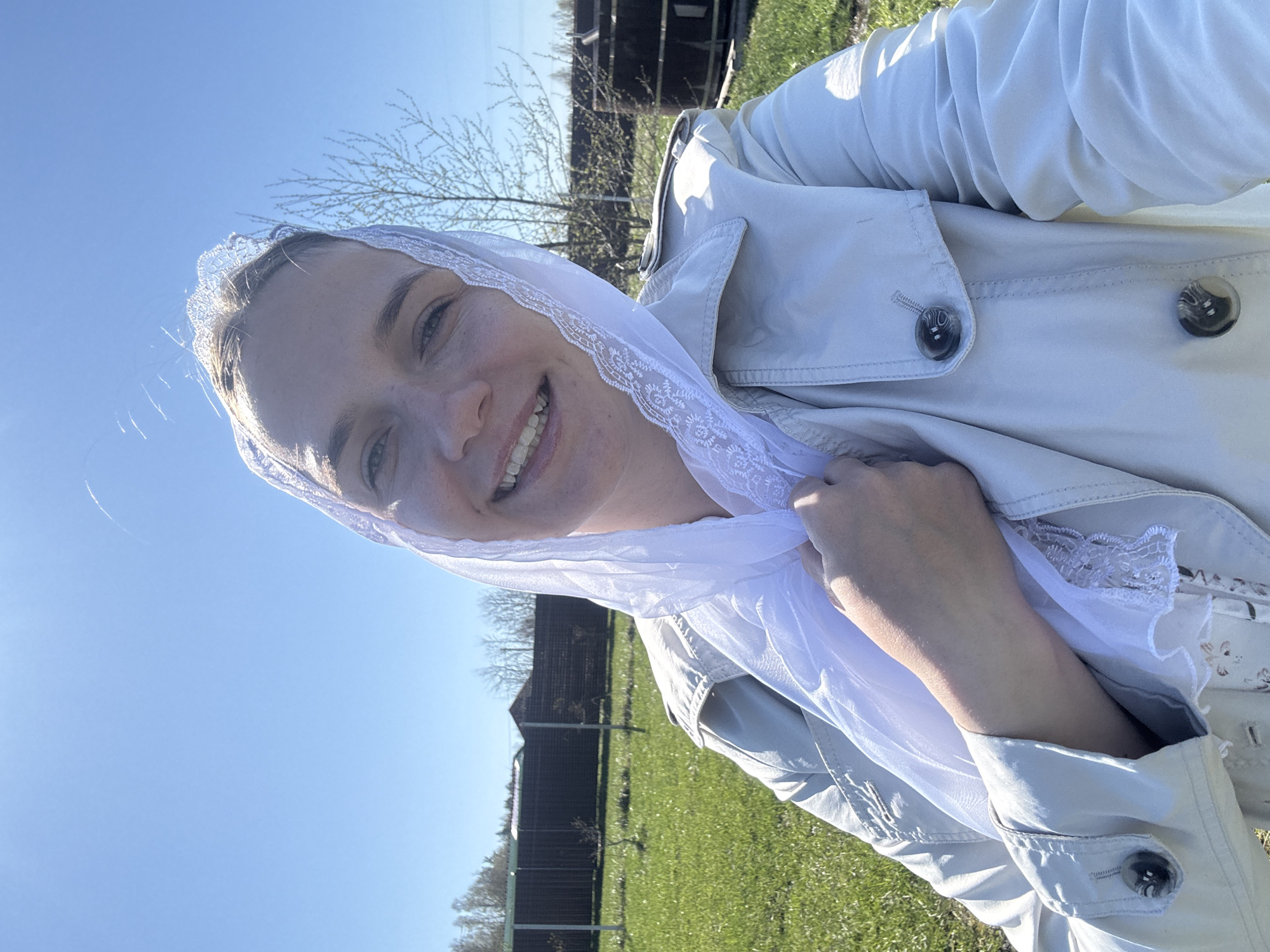 A woman is standing in an outdoor setting, likely in Раздолье, Russia. She is wearing a light-colored trench coat and a white lace veil. The environment consists of a grassy field, a fence, and trees. The sky is clear and blue.