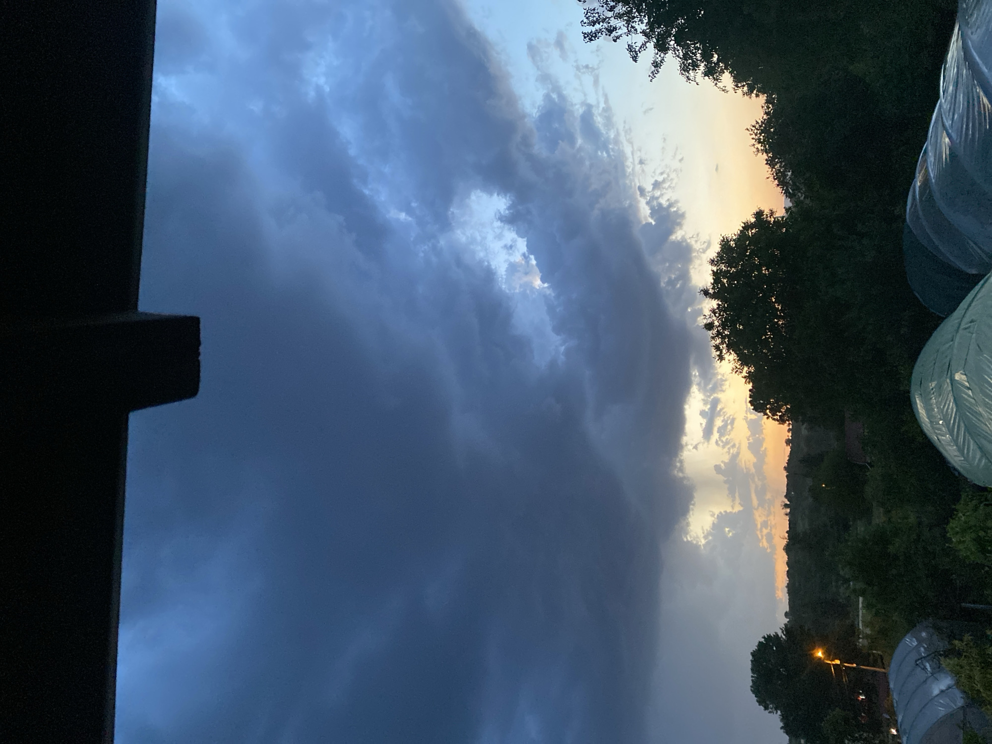 The image depicts a dusk or dawn sky partially obscured by dark storm clouds. The sky transitions from blue to a gradient of orange and yellow near the horizon. Silhouetted trees and a hill are visible along the lower right edge of the image, suggesting a rural or suburban setting. A covered structure, possibly a greenhouse, is partially visible in the upper right. A dark, unidentifiable structure with sharp edges is present in the left side.
Based on the location provided, the scene is likely in or near Konstantinovo, Bulgaria, though no landmarks specific to the city are discernible. The image contains no visible people or actions.