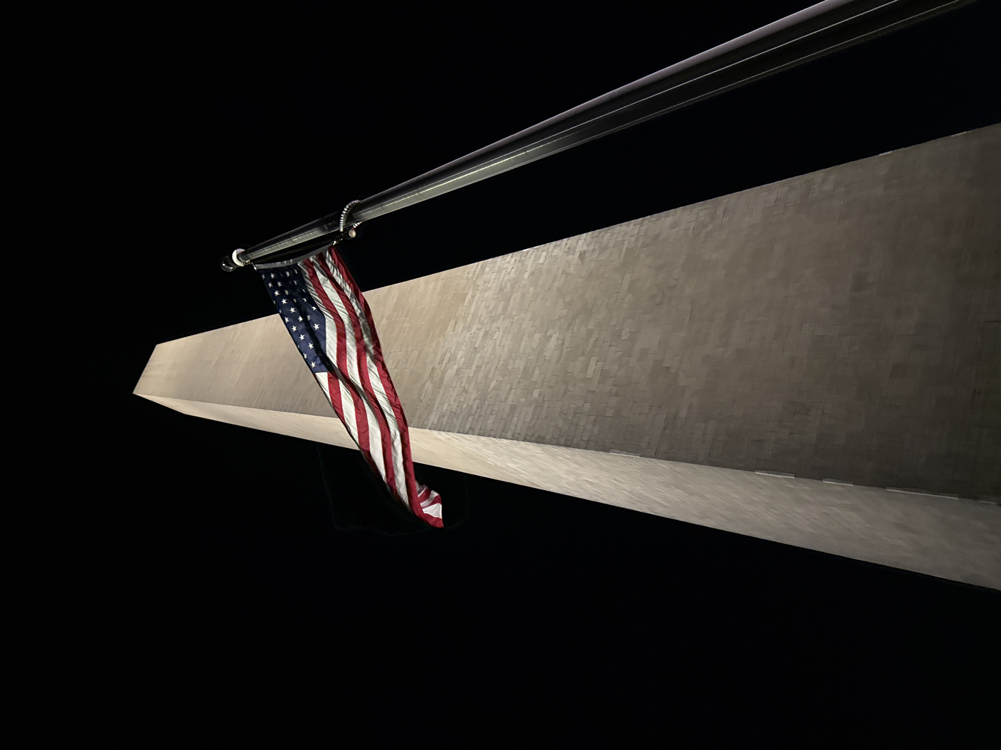 The image captures a low-angle view of the Washington Monument at night. An American flag hangs from a flagpole extending from the monument. The monument's facade is illuminated, revealing a textured surface. The sky is dark, suggesting nighttime. No people are visible.