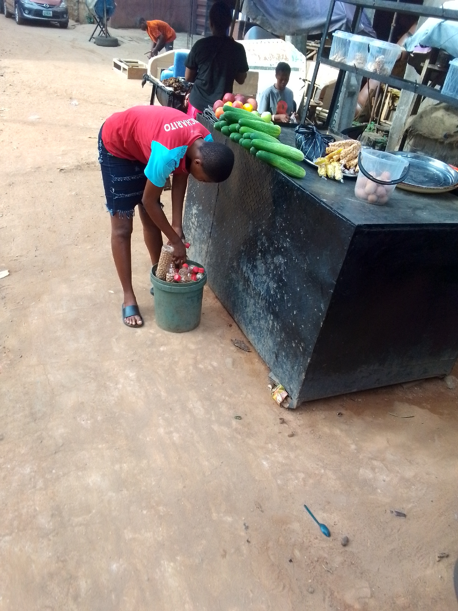 A young person in a red and blue shirt and denim shorts is leaning over a green bucket filled with items in small plastic bottles, likely packaged for sale. The bucket sits on the ground next to a dark, metal-clad stall where fruits, vegetables (primarily cucumbers), and packaged snacks are displayed. Other people are visible in the background, including one in a black shirt standing near the stall and another wearing orange working further back. The ground is sandy, and a spoon lies on the ground nearby. The background includes hints of a bustling outdoor environment with other stalls and a car parked nearby. The overall setting indicates a street market or roadside vending scene. The location is likely the city of Onitsha, Nigeria, based on the environmental cues and visual characteristics suggestive of the region.
