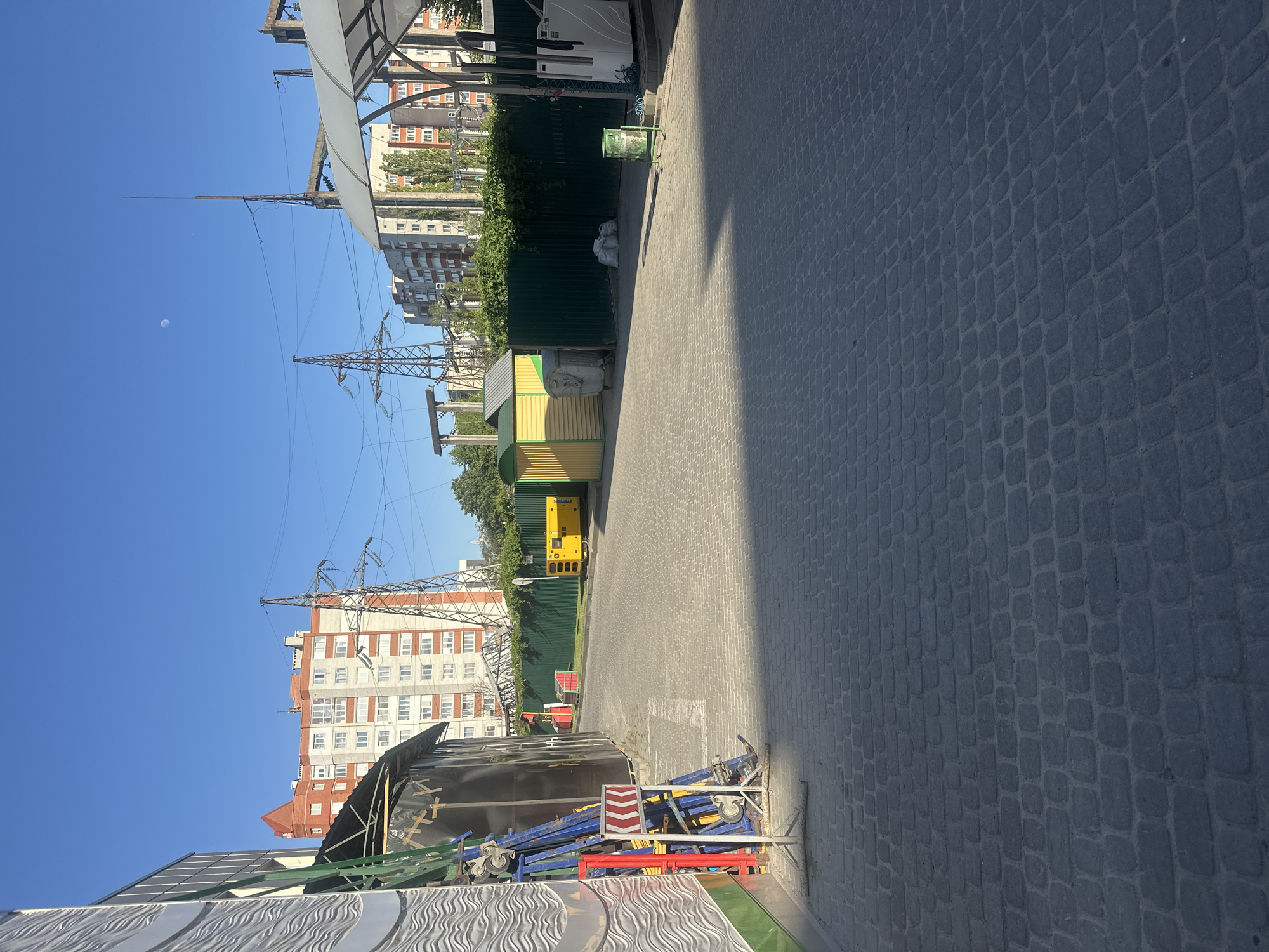 The image shows an alleyway paved with cobblestones, situated between buildings and a fence in Ternopil, Ukraine. A shadow bisects the alleyway. Overhead power lines and pylons are visible against a clear blue sky. A multi-story building with alternating red and white horizontal stripes is present in the background. Various objects, including a yellow generator, a green trash receptacle, and construction materials, are scattered along the alleyway. The moon is visible in the sky.