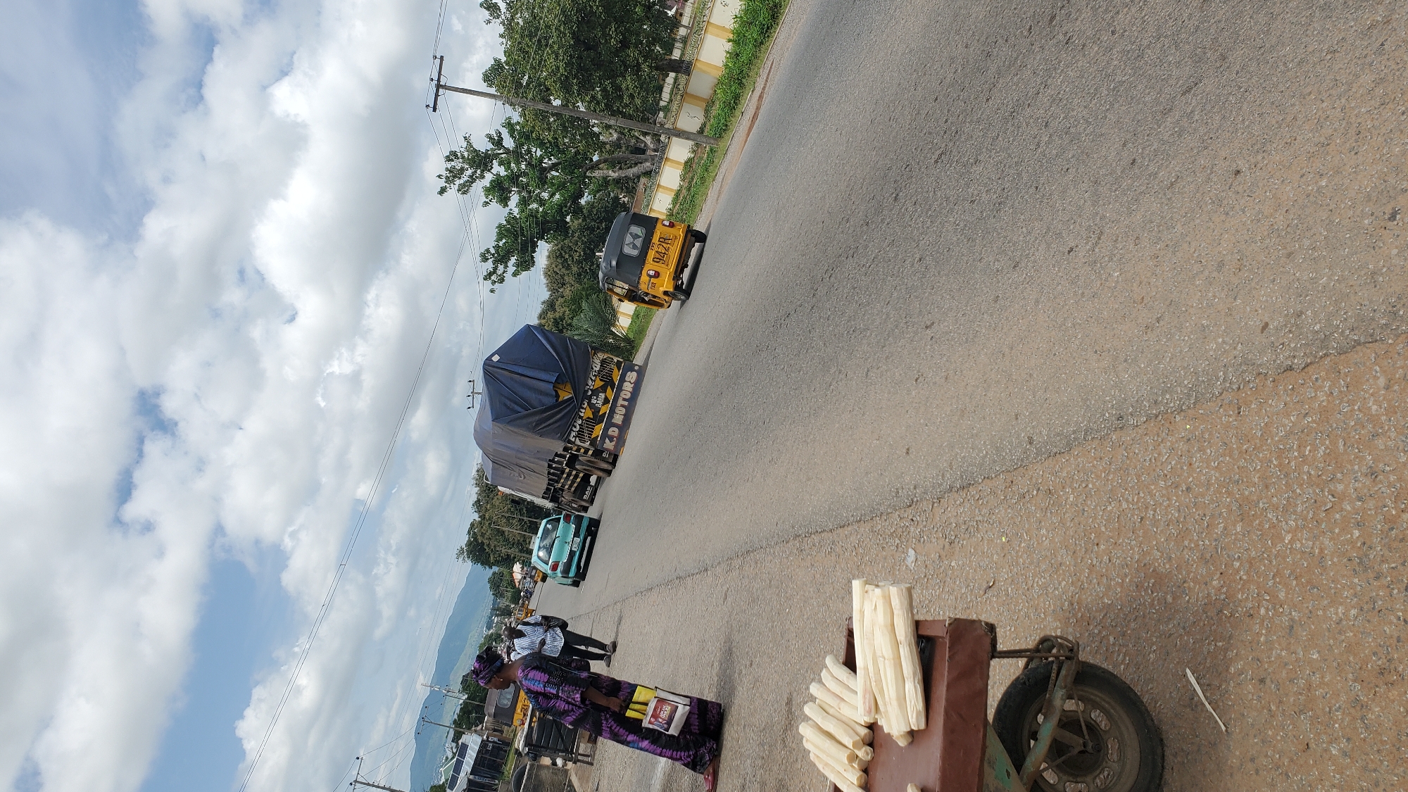 A street vendor is selling sugarcane sticks from a cart on the side of a road in Jalingo, Nigeria. The vendor, a woman wearing a patterned dress and headscarf, stands near the cart, holding shopping bags. In the background, a teal sedan is driving away from the camera, followed by a large truck covered with a blue tarp, labeled "K.D. MOTORS." A yellow three-wheeled auto-rickshaw is also visible on the road. The scene takes place under a cloudy sky with scattered trees and buildings visible in the distance. The road is paved, with a dirt shoulder.