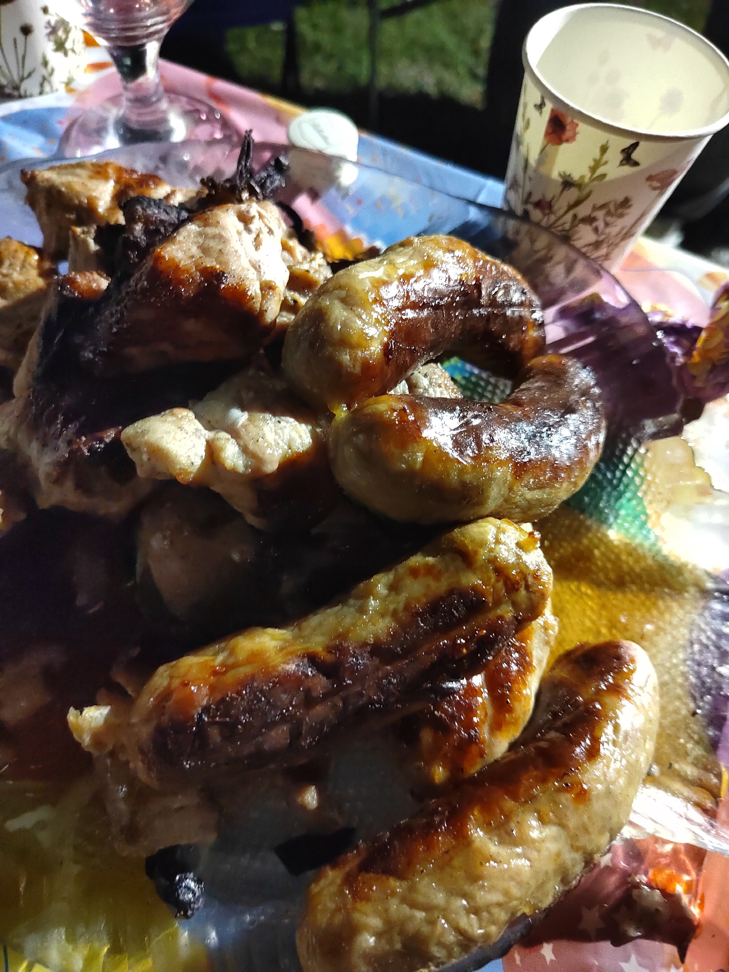 In a close-up, high-angle shot, a platter of grilled meats and sausages is presented on a table. The food, glistening with juices, appears to be the focus of the image, with a blurred background suggesting an outdoor setting. A decorative paper cup with floral motifs is visible to the right of the platter. The lighting suggests it is evening or night. The scene is outdoors, possibly at a picnic or gathering, in the city of Biysk, Russia.