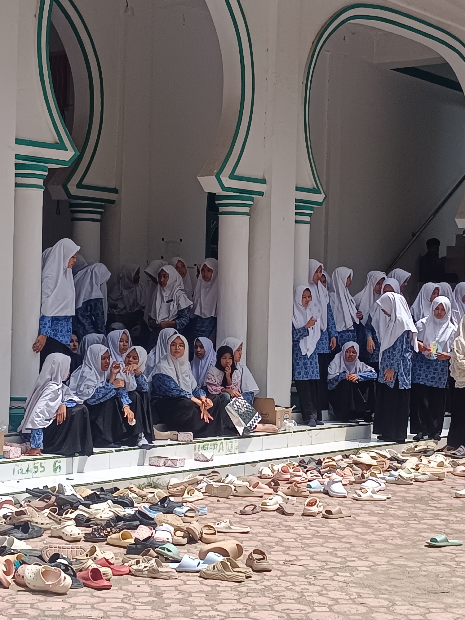 A group of approximately 20 young women wearing white headscarves and blue patterned tops with black skirts sit or stand on the steps of a building with white arches and green trim. The building appears to be a mosque or religious structure, evidenced by the large collection of shoes and sandals scattered on the brick-paved ground in front of the steps. A dark figure is visible in the background near a staircase. The location is likely Lhokseumawe, Indonesia, as indicated in the prompt. The women's actions suggest they are either waiting for or have just completed some religious activity or gathering. Several appear to be conversing. One girl sits with a patterned shopping bag. The day is sunny, casting shadows on the paved area.