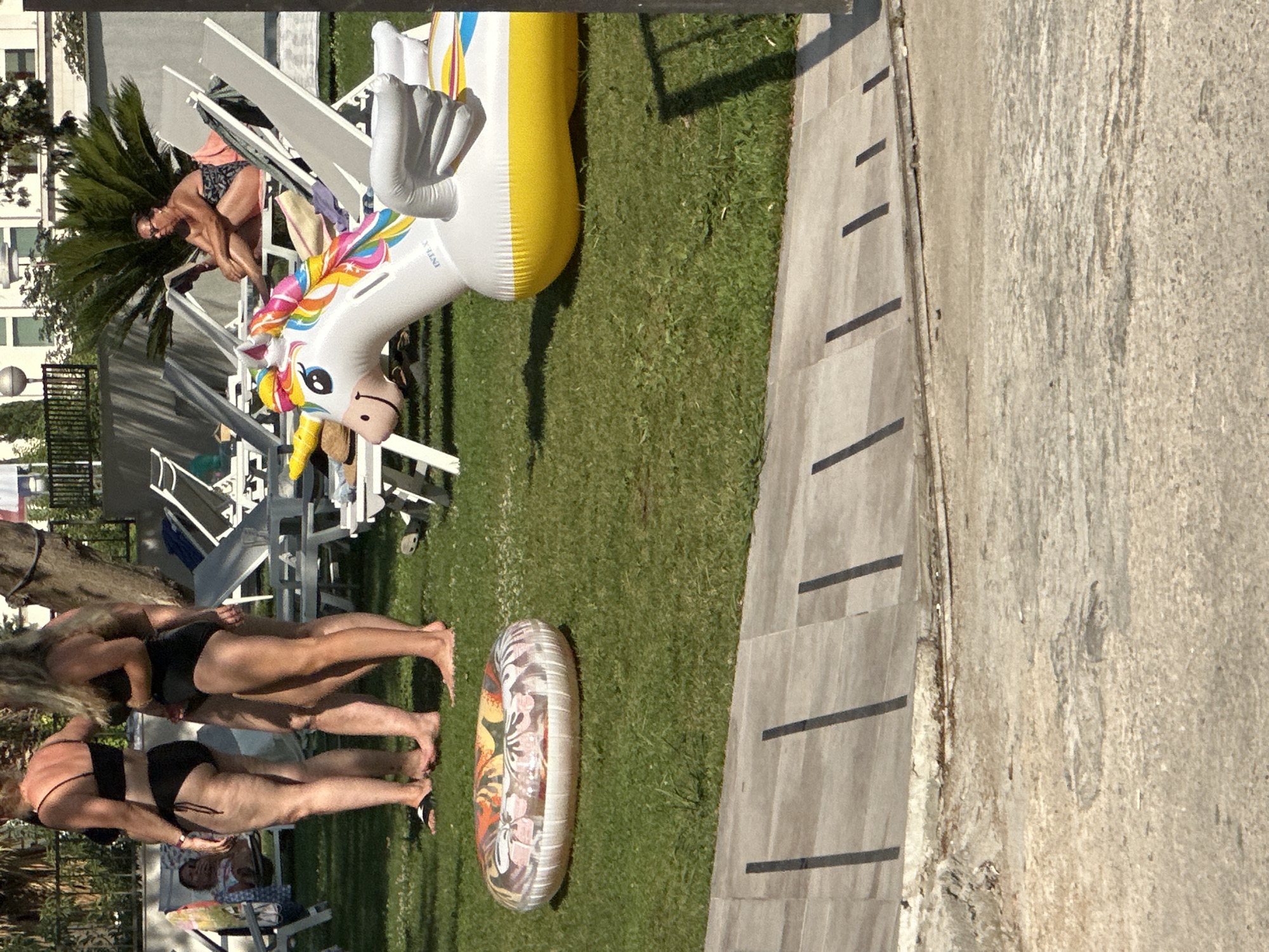 The image captures a sunny outdoor scene, likely at a resort or pool area in Bečići, Montenegro, with lush green grass and paved pathways. In the foreground, a large, inflatable unicorn with rainbow-colored mane and tail, and white wings, is partially visible on the grass, its yellow belly resting on the ground. To the left, a patterned inflatable ring lies on the grass. Three women are present: two are standing and dressed in black bikinis, their legs and torsos visible; the third woman is seated on a lounge chair in the background, wearing a patterned swimsuit and reading a book. Several white lounge chairs are scattered around the area, some occupied and some empty, suggesting a leisurely atmosphere. Palm trees and other vegetation are visible in the background, with buildings partially obscured. The scene is illuminated by natural sunlight, casting shadows on the ground.