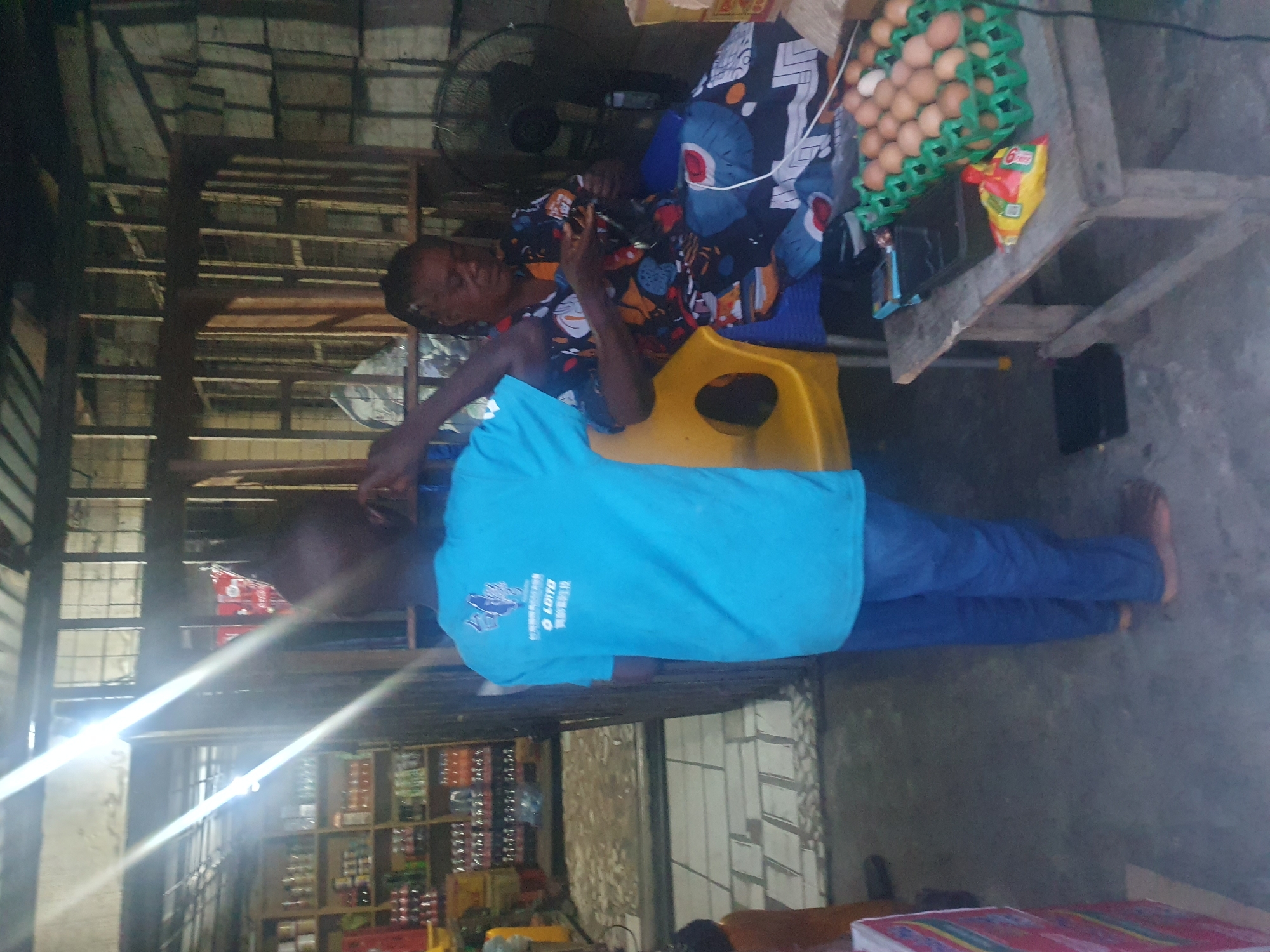 The image depicts two people in an indoor market setting in Lokoja, Nigeria. An adult female wearing a colorful dress and blue leggings sits on a yellow plastic chair, looking at a mobile phone held in her hand. A middle-aged man wearing a light blue t-shirt and blue jeans is seen standing next to her. There are shelves stocked with various goods, including canned food and drinks in the background. An egg carton is visible on a table near the seated woman. A standing fan is also visible in the background. The location appears to be a small, local market or store. The image is slightly rotated counter-clockwise.