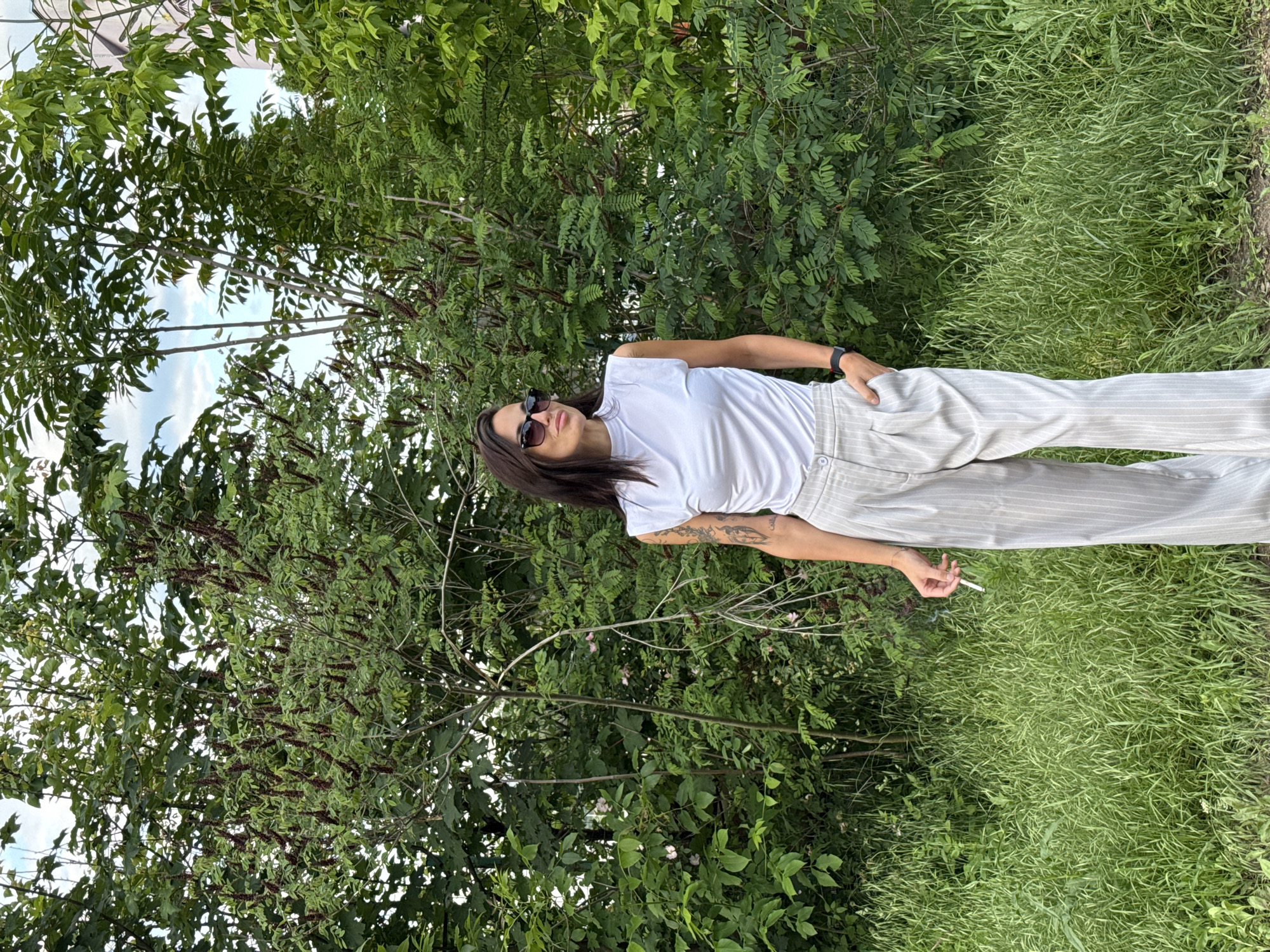 A young adult woman is standing in an outdoor, grassy area with dense green foliage and trees in the background. She is wearing sunglasses, a white T-shirt, and light-colored striped pants. She has dark hair and tattoos on her left arm. Her left hand is in her pocket, while her right hand is holding a lit cigarette. The location is speculated to be in Pleven, Bulgaria.