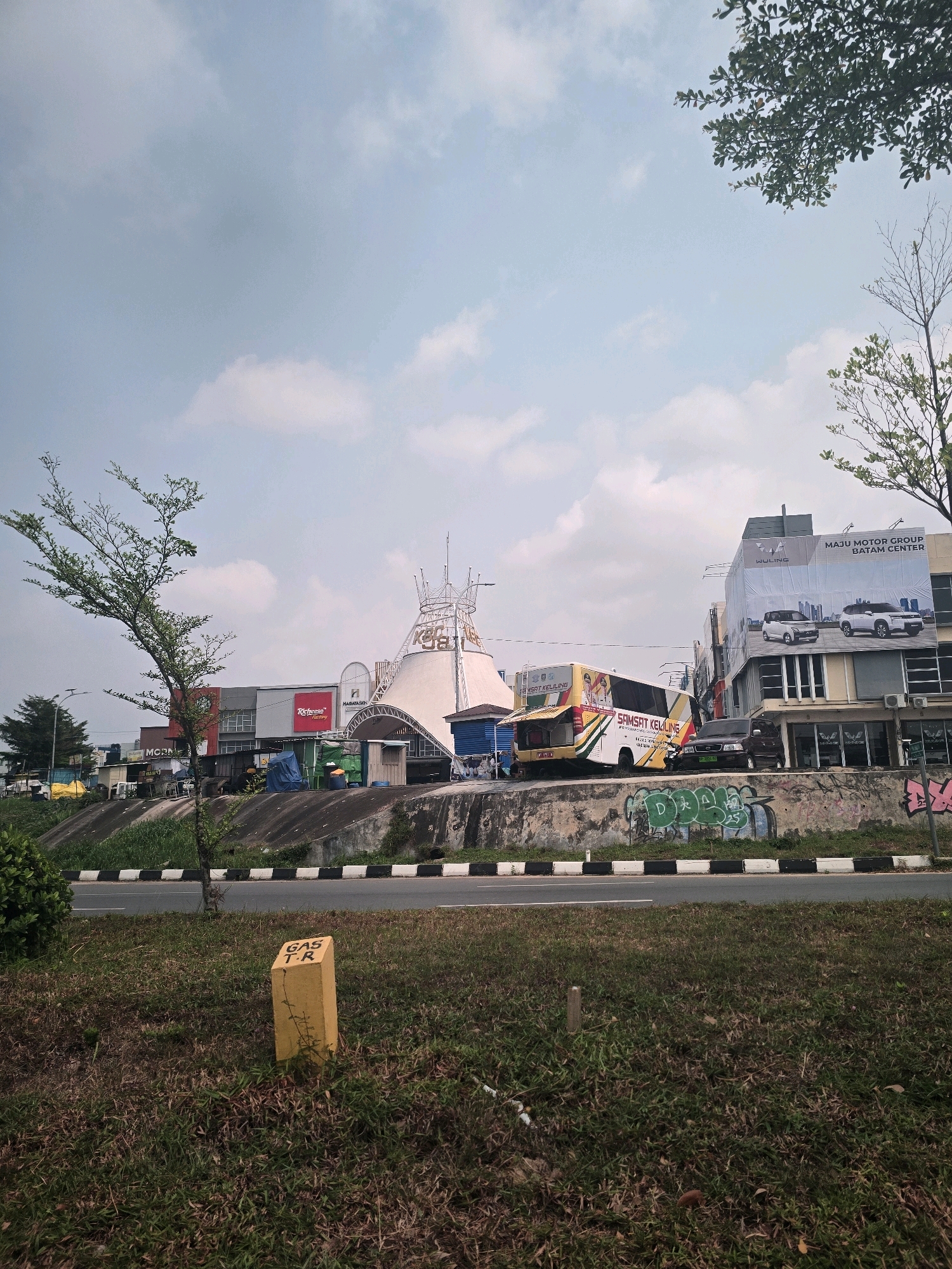 The image depicts a street scene in Batam, Indonesia, likely near a commercial area. In the background, a large, white, tent-like structure stands prominently, possibly a landmark or part of a public space. To the right of this structure, a multi-story building displays a large advertisement for "MAJU MOTOR GROUP BATAM CENTER" featuring images of cars. A yellow and white bus with "SAMSAT KELILING" written on its side is parked near the building, suggesting it's a mobile service unit. Several other businesses with signs like "MCRN" and "Richeese Factory" are visible along the street. The area is bordered by a concrete retaining wall covered in graffiti, with a road in the foreground. A small yellow marker with "GAS T.R" inscribed on it is positioned in the grassy median strip. Trees and foliage are scattered throughout the scene, with a large leafy tree in the foreground on the left and branches of another tree visible at the top right. The sky is partly cloudy, indicating daytime.