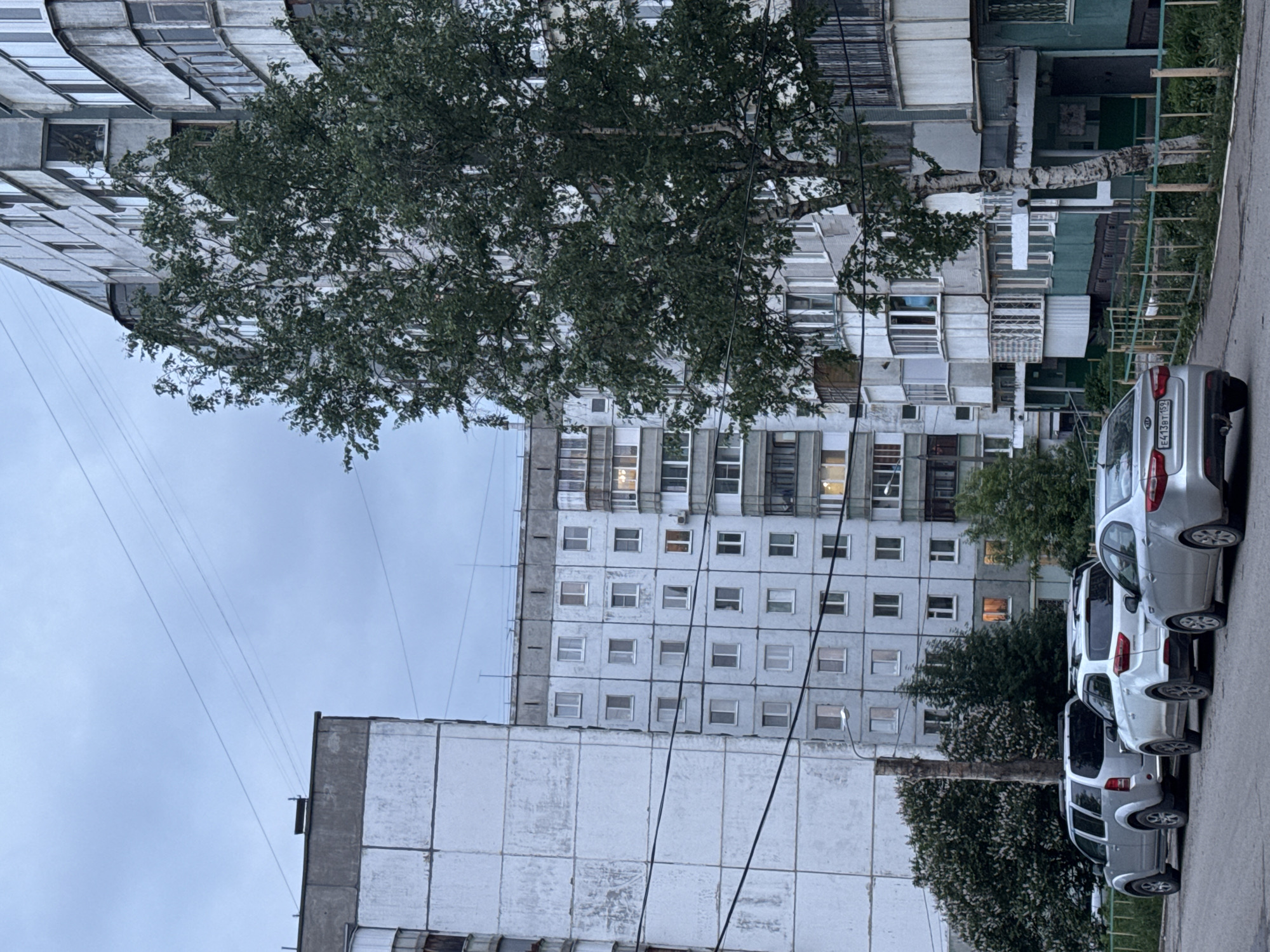The image shows a street in Perm, Russia, with several multi-story apartment buildings. The buildings appear to be of Soviet-era construction, with plain, repetitive facades. A large tree partially obscures the view of one of the buildings. Several parked cars line the right side of the street. The sky is overcast. There are power lines running overhead.