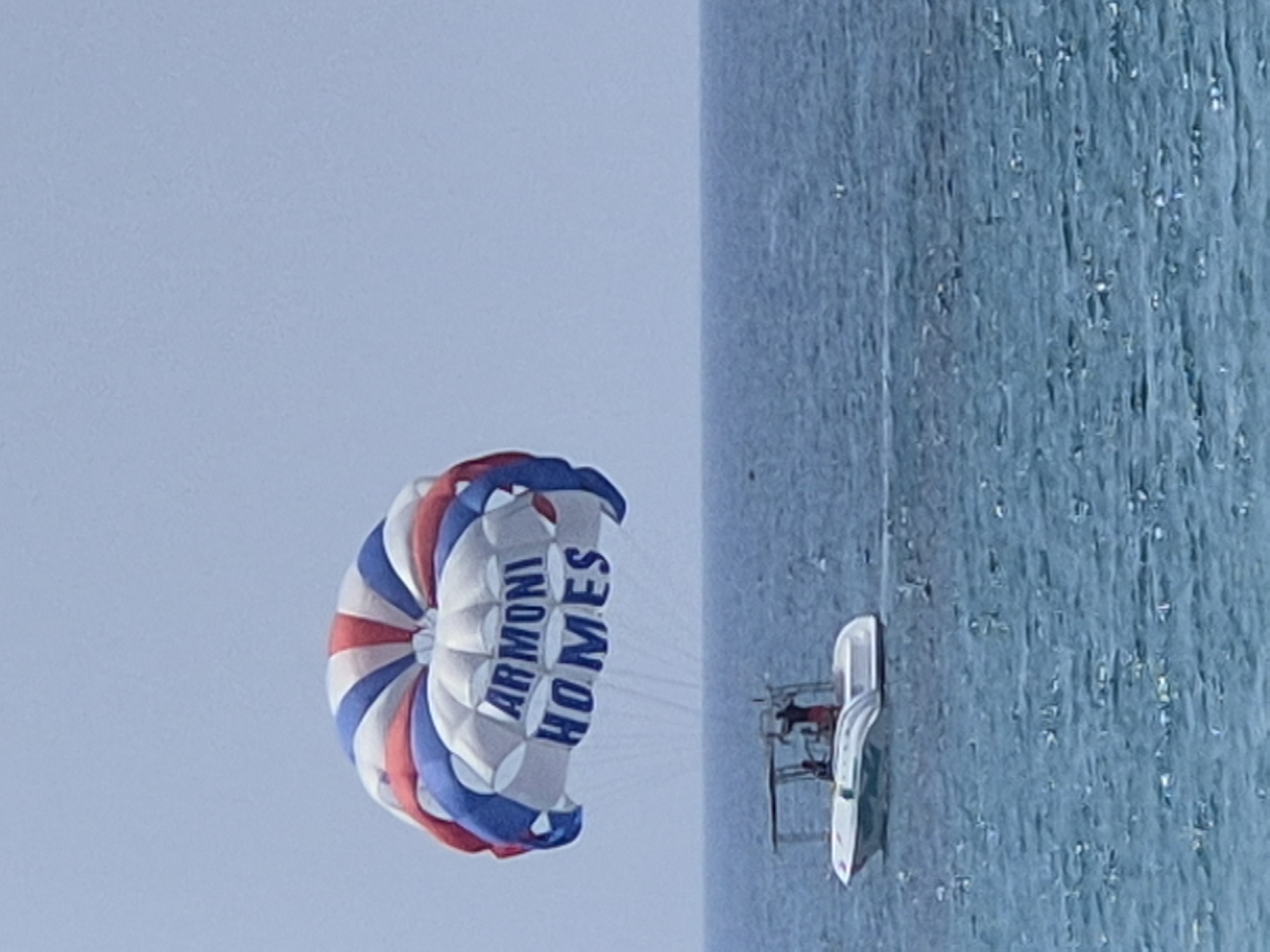 A parasail with the words "ARMONI HOMES" printed on it is being pulled by a boat across the blue waters off the coast of Alanya, Turkey. The parasail is red, white, and blue, and it is high in the sky. The boat is white and has a canopy. There are two people visible in the boat. The sky is clear and blue. The image appears to have been taken on a sunny day.