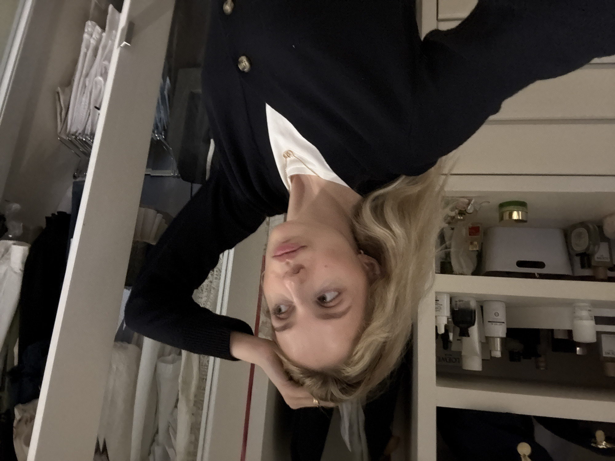 A young woman with blonde hair is standing in front of an open closet. She is wearing a black cardigan over a white t-shirt and a gold necklace. Her right hand is resting on her head. The closet shelves are filled with various toiletries and clothing. The lighting is dim. The image does not provide any information about the location of Mexico City, Mexico.
