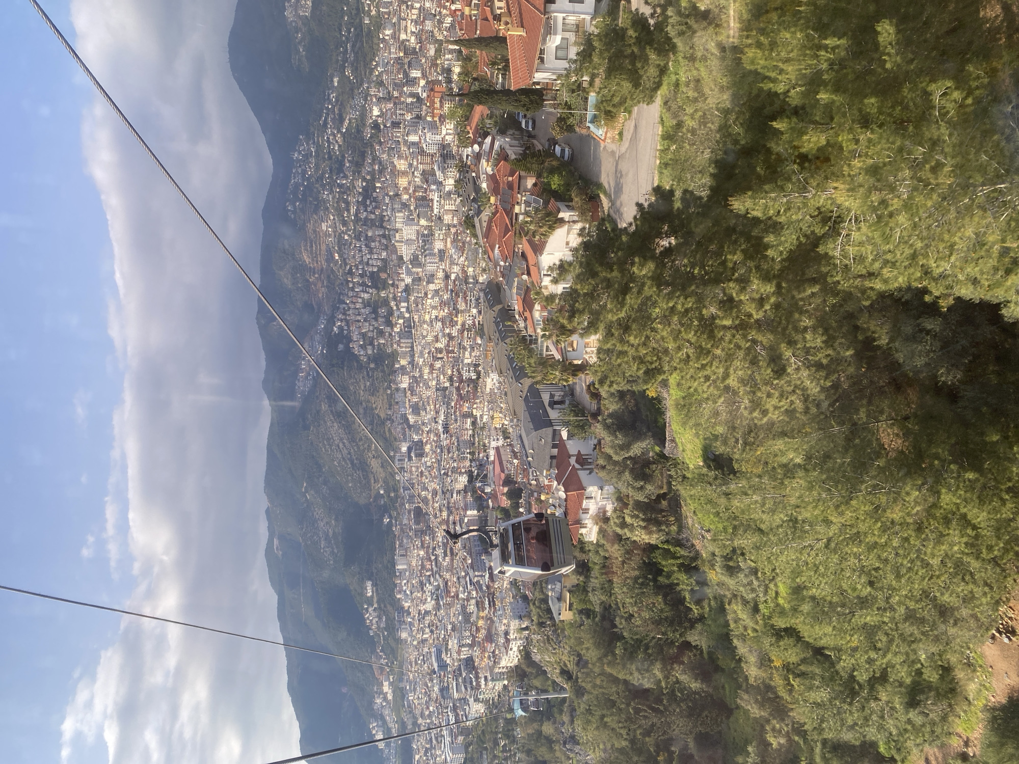 The image depicts a cable car descending towards the city of Alanya, Turkey. The densely populated city stretches across the landscape, with buildings primarily colored white and beige. Red-tiled roofs are interspersed among the structures. Mountains rise behind the city, and the sky is partially covered by clouds. The foreground of the image features the cable car, with visible cables extending from the car towards the ground and up towards the mountaintop. The interior of the cable car is visible, but the occupants are not clearly discernible. To the right of the cable car, there are lush green trees and vegetation. A road runs alongside the edge of the city, adjacent to the tree line.