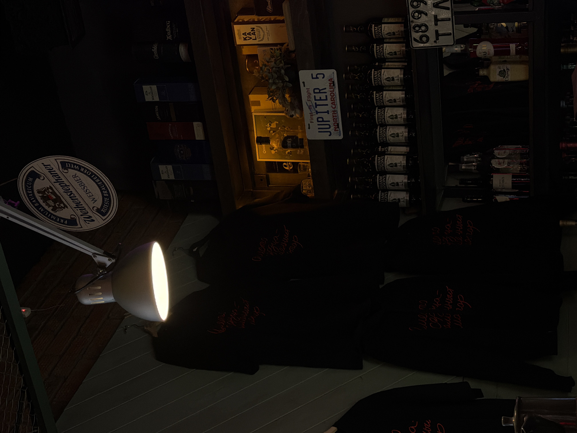 This dimly lit scene appears to be a retail or bar environment in an unspecified location. The photograph is composed with an angled perspective.
In the foreground, several black t-shirts or hoodies with embroidered red script (possibly Russian Cyrillic) are partially visible. A large, industrial-style lamp casts a beam of light onto the clothing.
Behind the clothing is a display of alcoholic beverages on multiple shelves, including brands such as Macallan, Glenfiddich, Ardbeg, and Jameson. There are multiple bottles of what appears to be "William Lawson's" whisky. A decorative arrangement of flowers and foliage is positioned among the bottles. A novelty license plate reading "JUPITER 5 NORTH CAROLINA First in Flight" is noticeable.
A coaster or sign displaying the "Weihenstephaner" brand is positioned on the left side.
The angle of the image obscures the full view of the room.