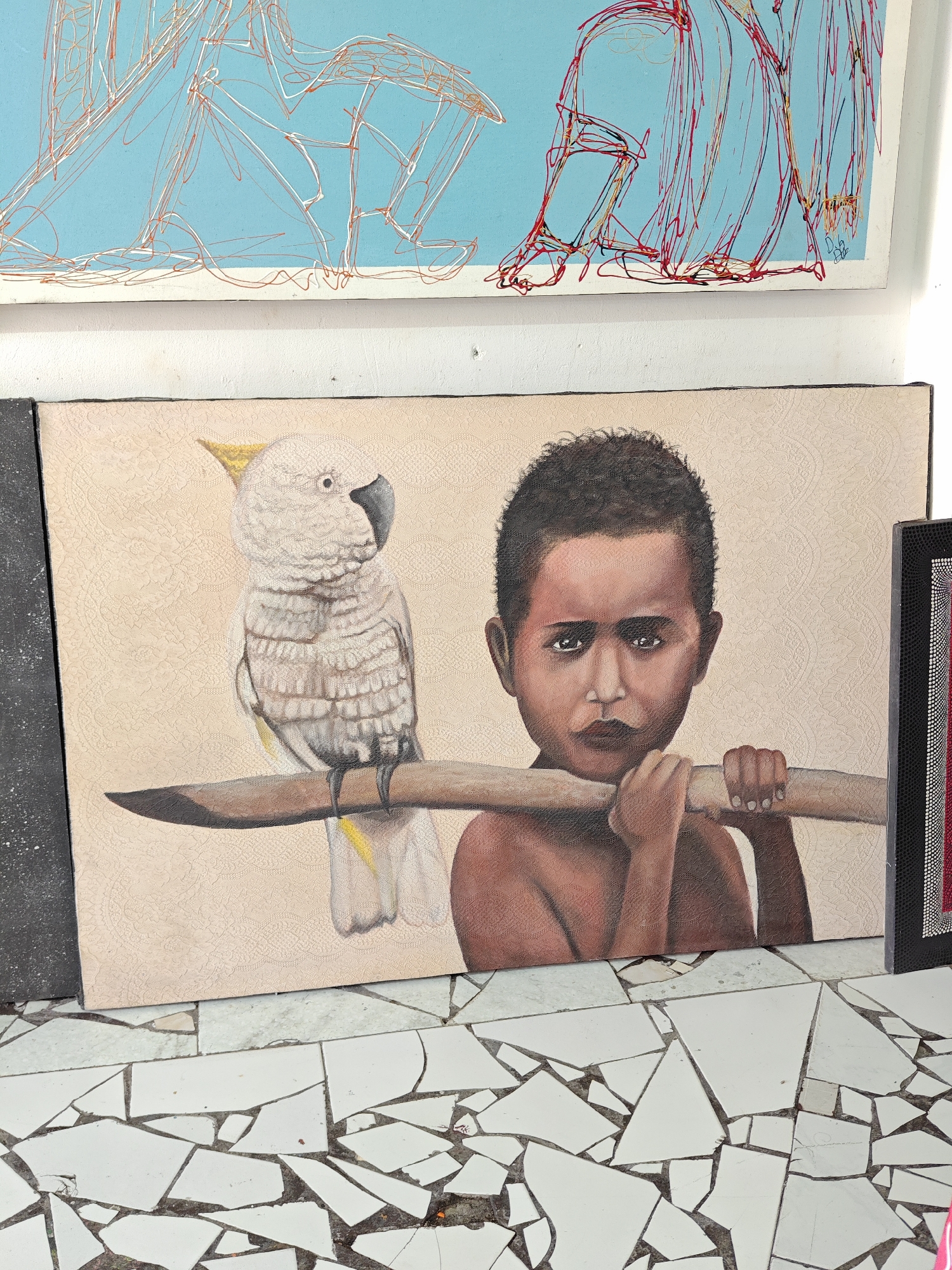An image showing an assortment of artwork and flooring, taken in Denpasar, Indonesia. A painting of a young boy carrying a branch with a white cockatoo perched on it is prominently displayed, positioned beneath a blue and white painting with linear red accents of two figures. The bottom portion of the image shows the flooring which appears to be made of white tiles.