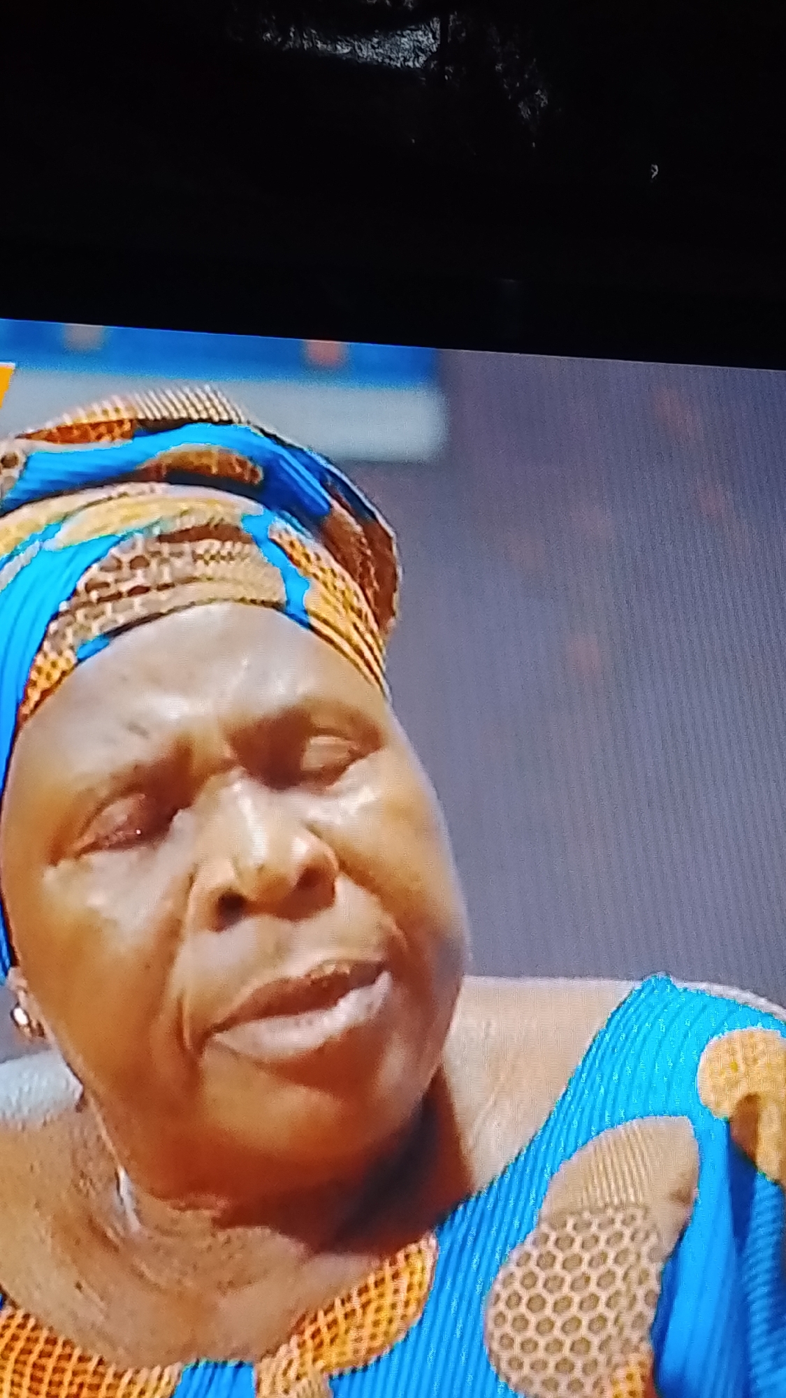 The image shows a close-up of an older Black woman wearing a blue and orange patterned headwrap and a matching blue garment with orange circular patterns. Her eyes are closed, and her mouth is slightly open, suggesting she might be speaking or singing. The lighting is somewhat dim, with the focus on her face and upper body. The background is blurred and appears to be an indoor setting, possibly a room. There is no visible indication of the specific location within Lokoja, Nigeria.