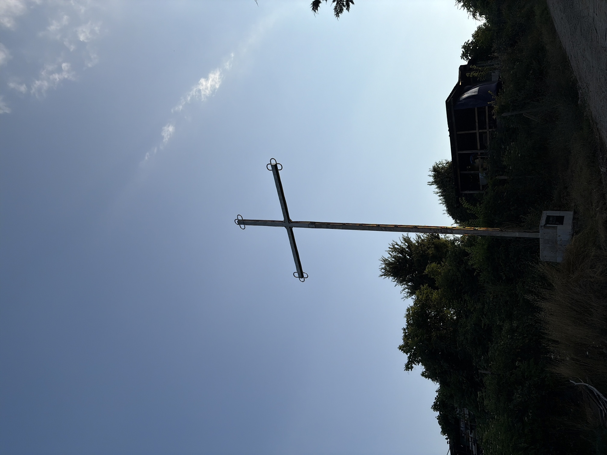 The image depicts an outdoor scene at a low angle, with a clear blue sky dominating the upper portion. In the center, a tall, metallic cross stands prominently, silhouetted against the sky. The cross has a simple design with two curved elements at the ends of the horizontal beam. It is mounted on a concrete base.
In the lower half of the image, dense green foliage and trees form a dark, natural backdrop. Behind the trees, a structure that appears to be a makeshift shelter or building with a wooden frame and a blue tarp is visible. A dirt path or road curves from the lower right corner into the scene. The overall impression is one of a somewhat rural or undeveloped area. The image does not contain any people or visible actions.