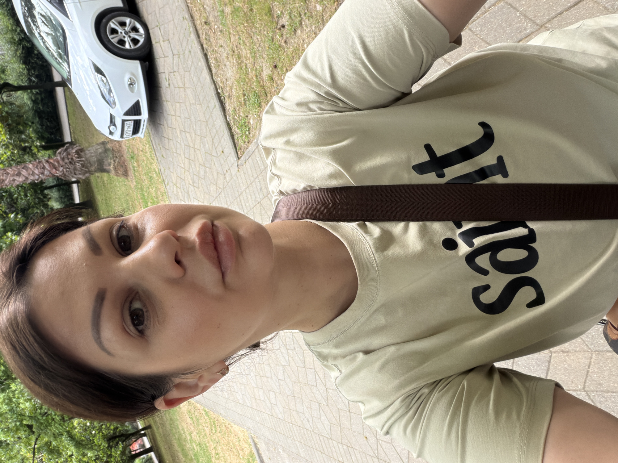 A young woman with short, dark hair and brown eyes takes a selfie outdoors. She is wearing a light green t-shirt with the word "saint" printed in black on the front, and a brown strap across her chest. Her face is turned towards the camera, and her lips are slightly parted. In the background, a white car is parked on a paved area, with grass and trees visible beyond. The scene appears to be a park or residential area in Sochi, Russia.