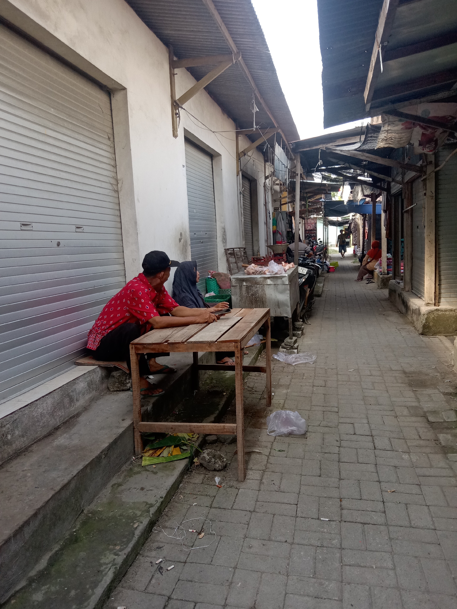 The image depicts a street scene in Klepek, Indonesia, characterized by a narrow alleyway lined with closed market stalls. Two individuals are seated at a wooden table in the foreground on the left side of the alley. The person on the left, a male wearing a red patterned shirt and a black cap, is looking down at his phone. To his right sits a person wearing a dark headscarf, partially obscured. The alleyway is paved with interlocking bricks, and the stalls on either side have corrugated metal roofs and roll-up metal doors. In the background, other market stalls are visible, some open with merchandise displayed, and a few people are walking through the alley. A motorcycle is parked to the right of a stainless steel counter displaying what appears to be raw meat. The overall environment appears to be a bustling marketplace that is either beginning to open or closing down for the day, given the closed shutters of some stalls. The lighting suggests it is daytime, possibly overcast.