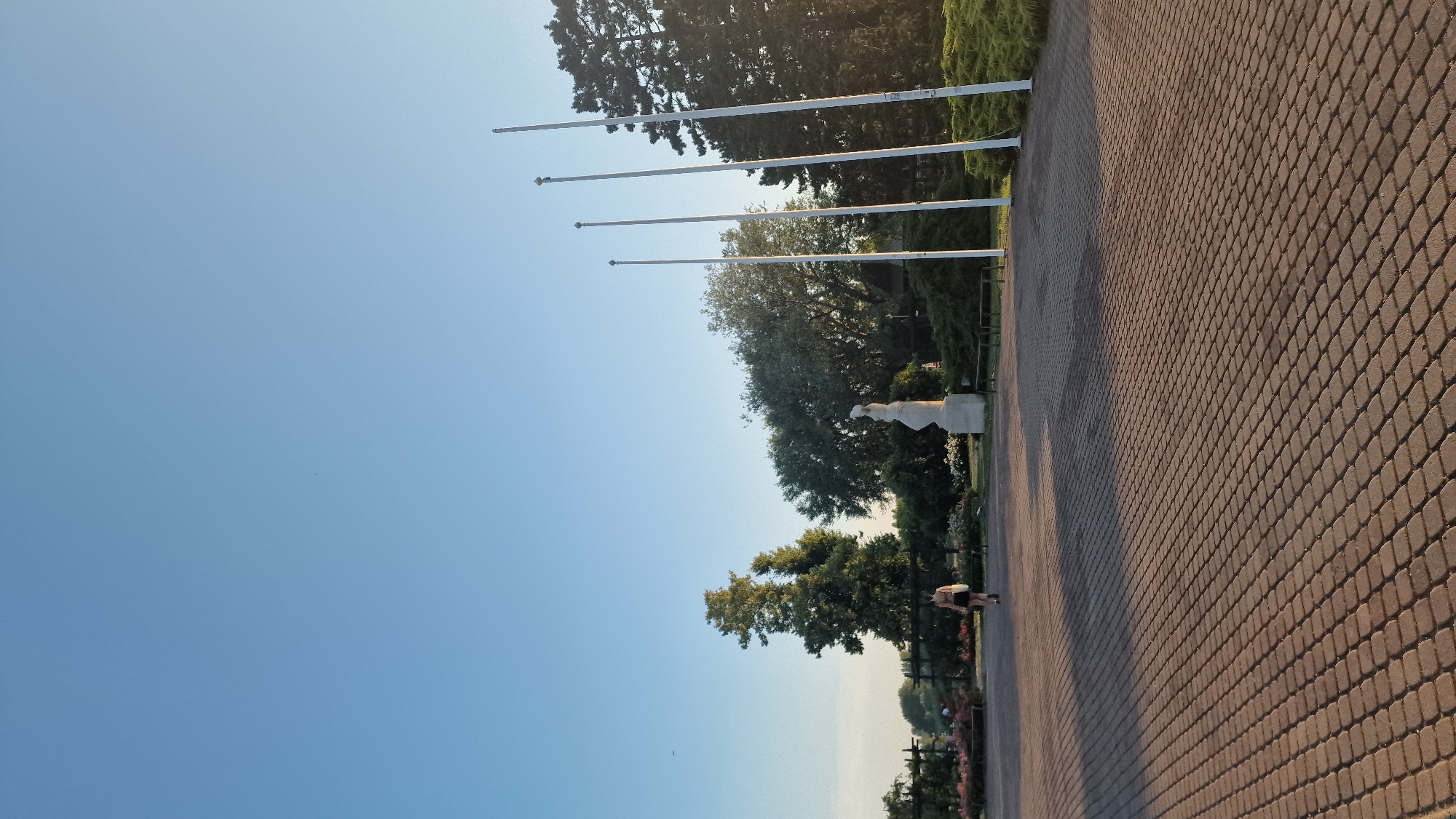 A woman walks across a paved plaza in front of a statue and several flagpoles. The scene is set in a park with trees and flowerbeds in the background. The sky is clear and blue, and the sun casts long shadows across the plaza. This image was taken in Keszthely, Hungary, specifically in the area surrounding the Helikon Palace Museum.